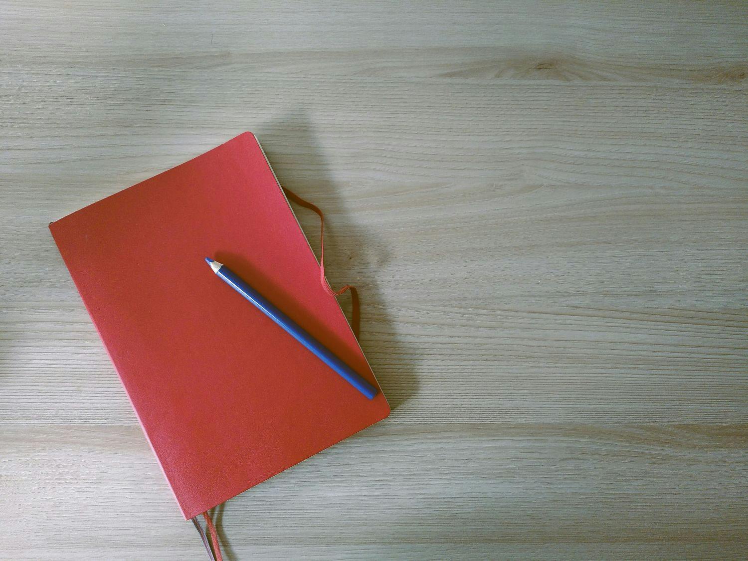 Rotes Notizbuch liegt auf dem Tisch, auf dem Notizbuch liegt ein blauer Farbstift.