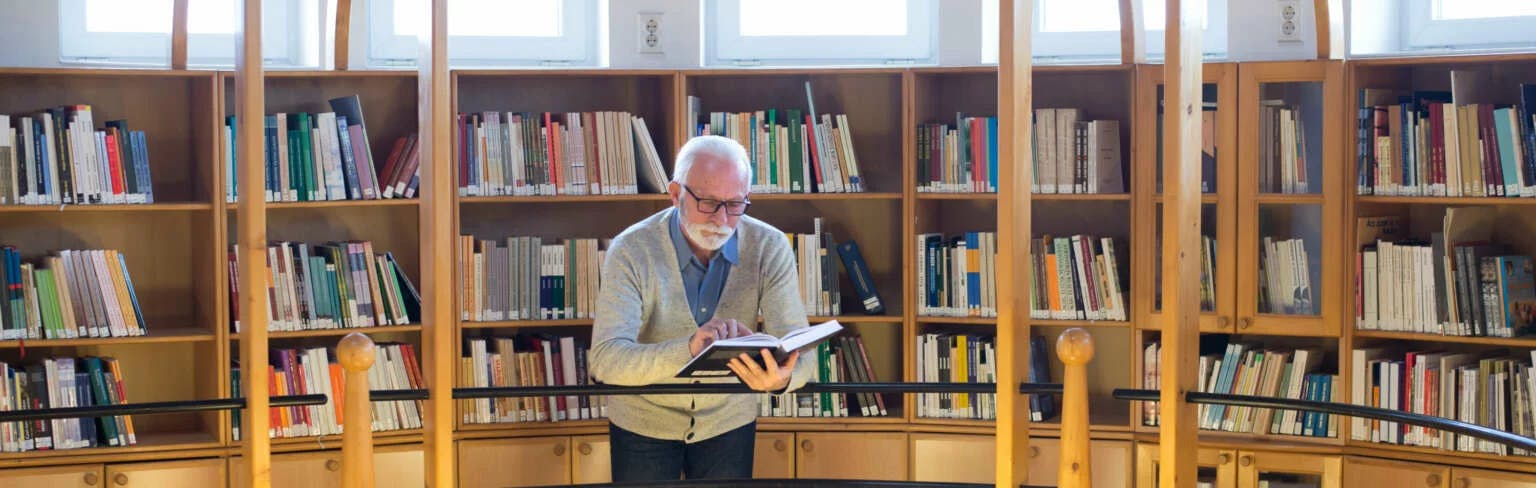 Älterer Herr mit weißem Haar und weißem Bart steht vor einem Buchregal und liest in einem Buch
