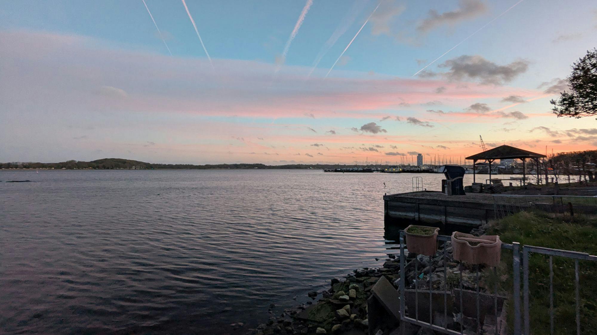 Ein Blick über die Schlei bei Schleswig in der Abenddämmerung. Die Wolken werden von der untergehenden Sonne rötlich gefärbt. Am Horizont jenseits des Wassers ist der Hügel zu erkennen, in dessen Nähe sich die alte Wikingersiedlung Haithabu befindet.