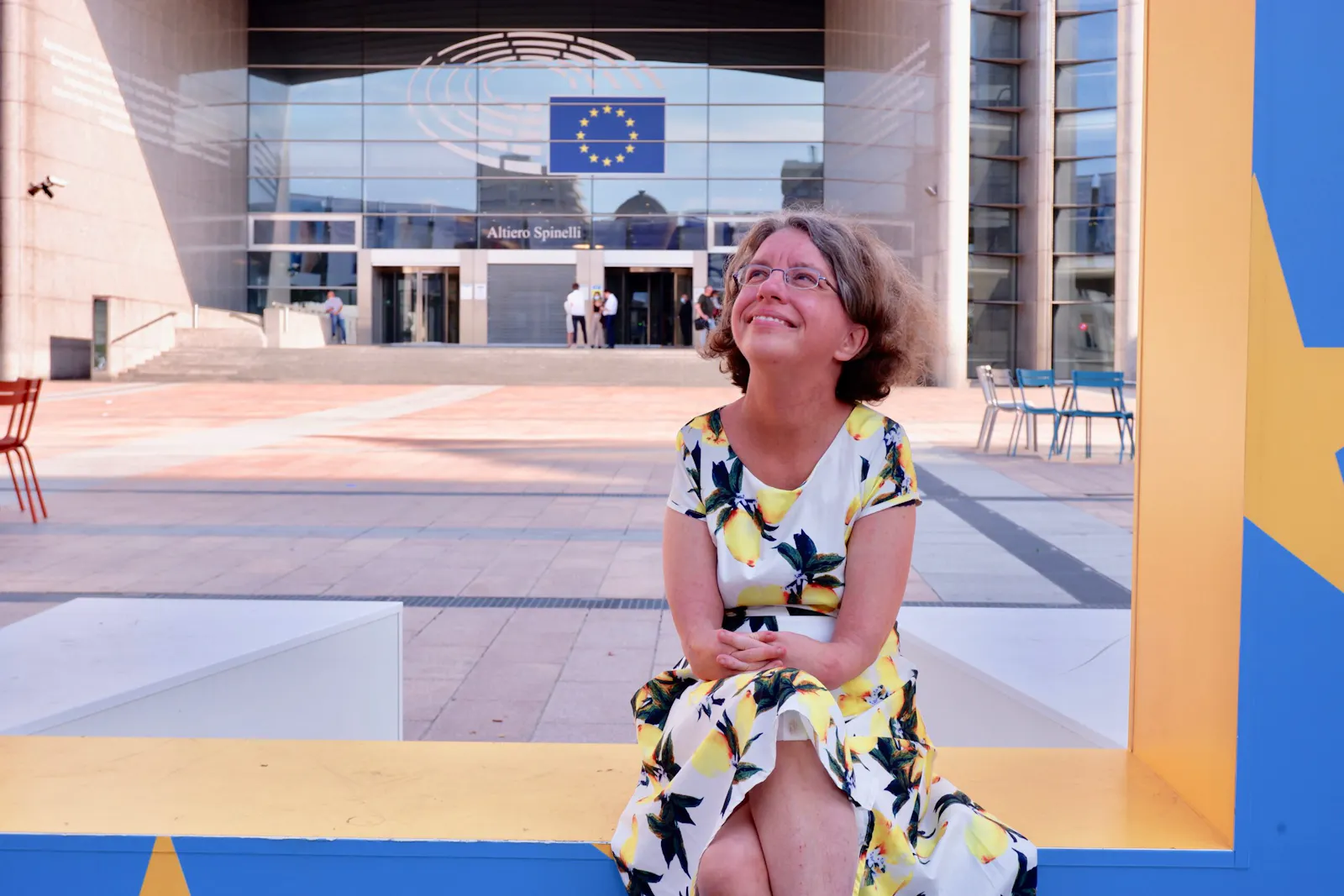 Katrin Langensiepen sitzt in einem Sommerkleid mit Zitronenmotiv vor dem Europäischen Parlament und schaut lächelnd nach rechts oben aus dem Bild heraus.