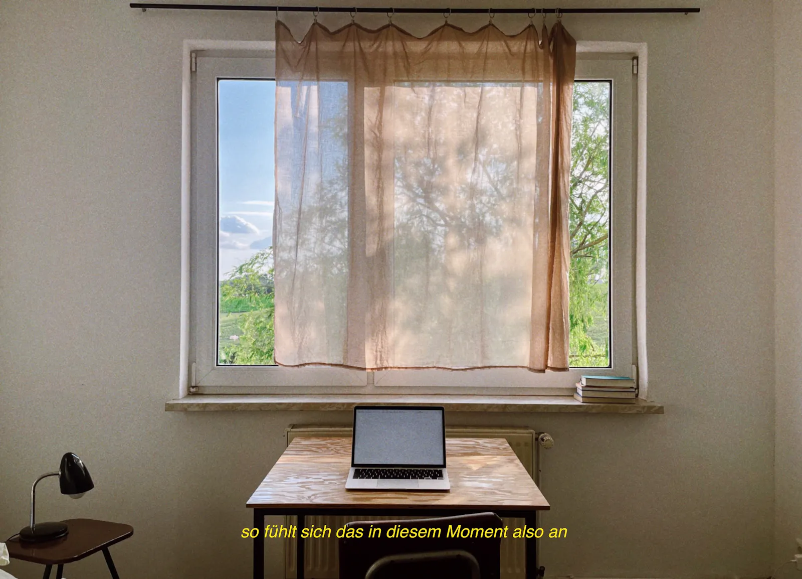 Ein Schreibtisch vor einem Fenster, darauf steht ein aufgeklappter Laptop. Unten in gelber Schrift "so fühlt sich das in diesem Moment also an"