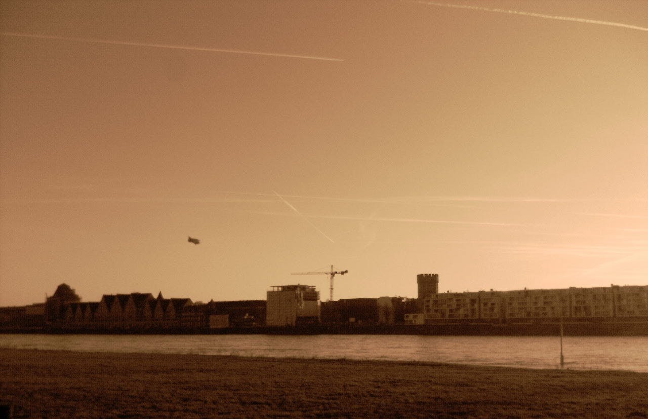 Ein Bild in Sepia, Blick auf den Rhein, auf der anderen Flusseite sind Häuser und ein Baukran zu sehen. In der Luft fliegt ein Zeppelin.