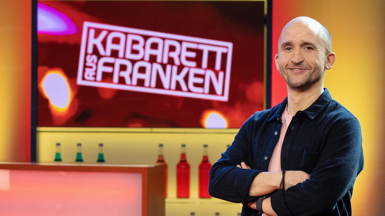 Ich stehe vor der Kulisse von Kabarett aus Franken: im Regal einige bunte Flaschen, dahinter das Logo der Sendung.
