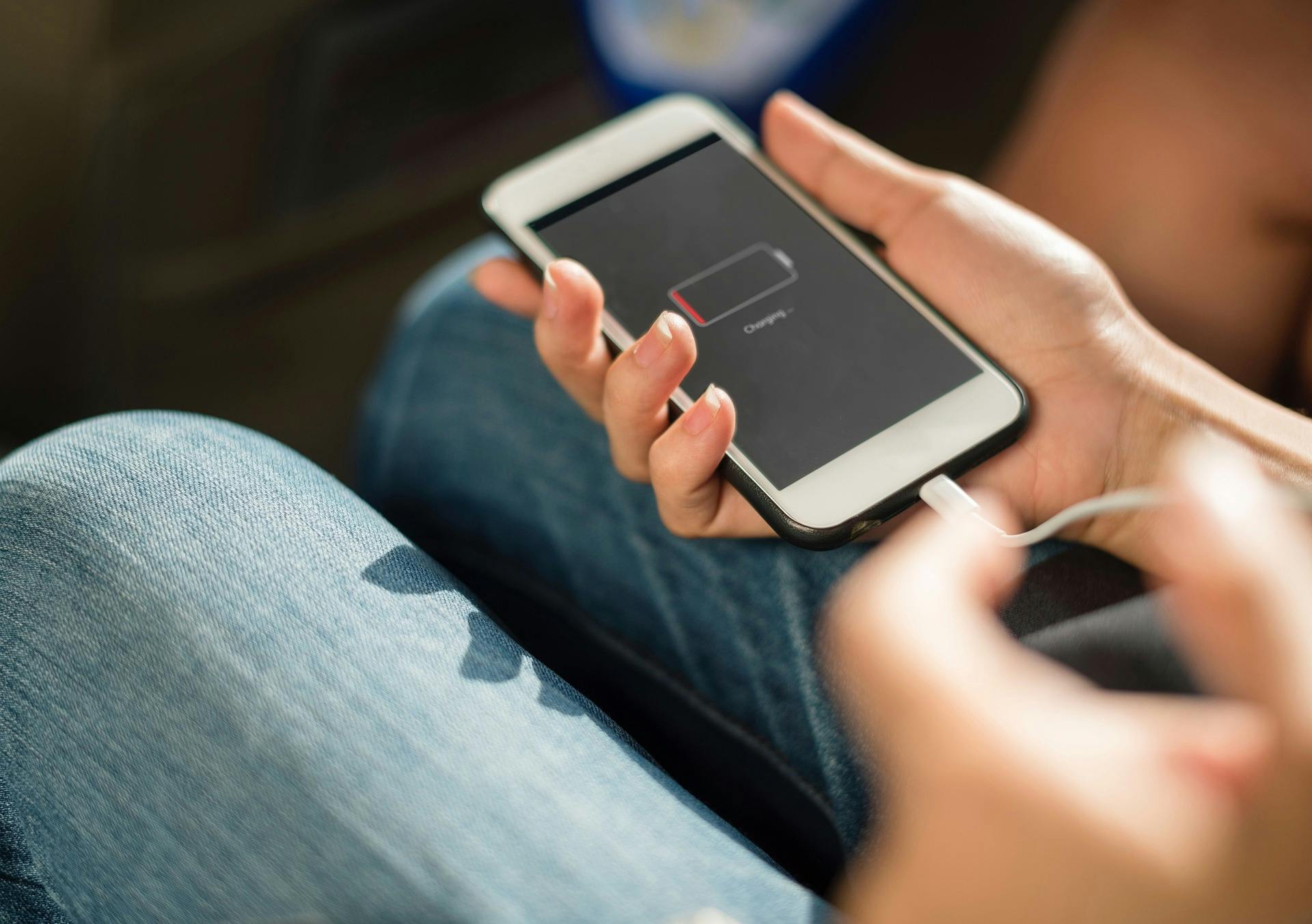 Im Vordergrund ein Handy, mit einem leeren Akku, im Hintregrund eine sitzende Person, die das Handy in der Hand hält und das Handy auflädt