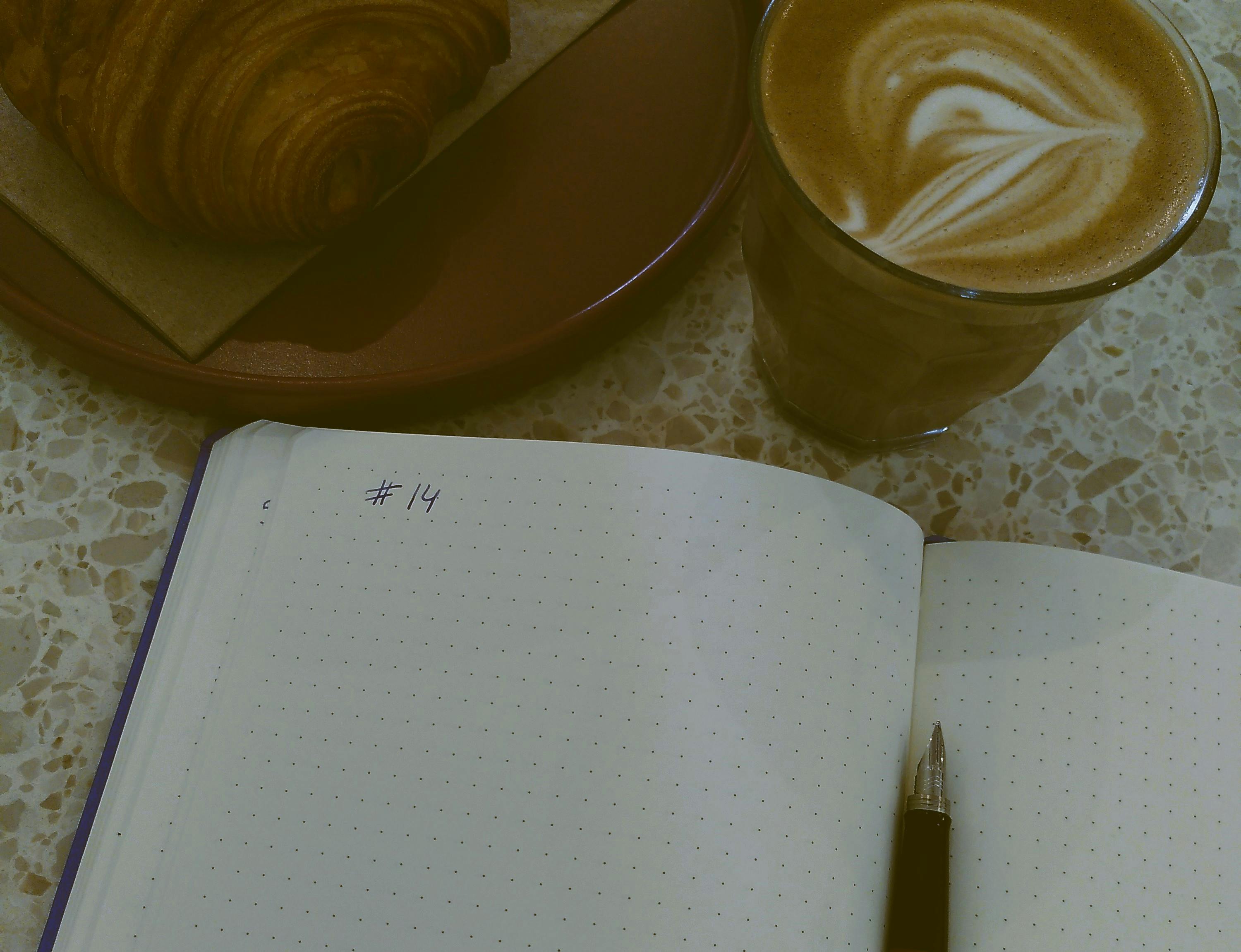 Ein offenes Notizbuch mit einem Füller in der Mitte, auf der linken Seite steht #14. Oben links im Bild liegt ein Schokocroissant auf einem Teller, daneben steht ein Cappuccino.