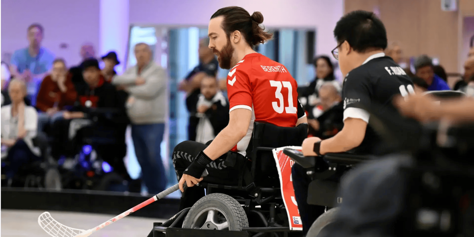 Zwei Power-Hockey Spieler auf dem Feld in Aktion, im Hintergrund Zuschauer*innen.