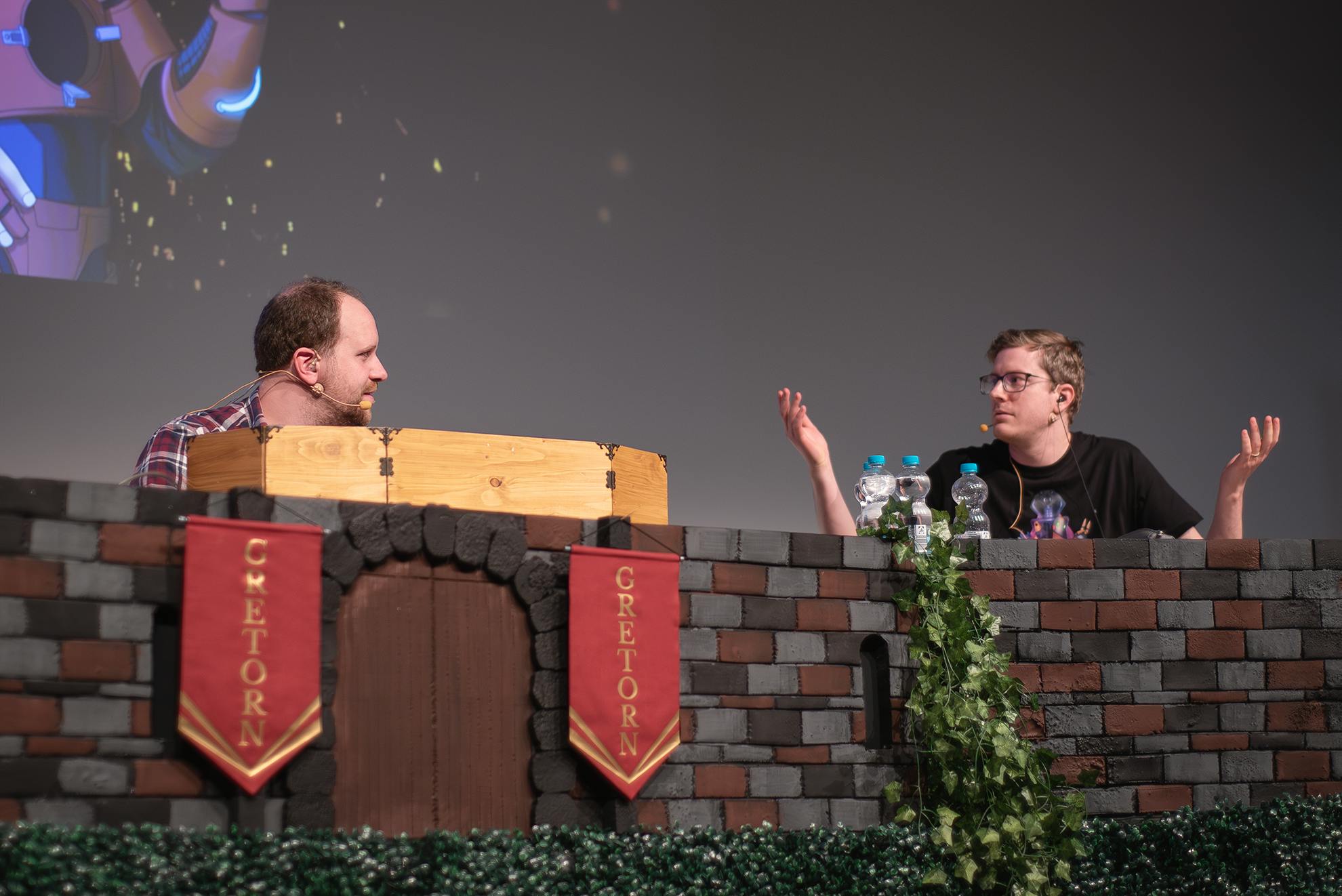 Alex und Tim sitzen während der Liveshow hinter einer Bühnendeko, die Mauerwerk darstellt.