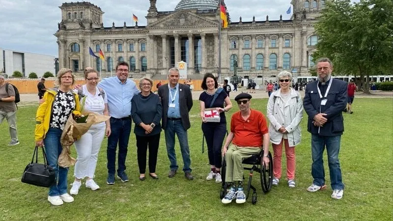 Das Bild zeigt eine Gruppe von neun Personen, die gemeinsam vor dem Reichstagsgebäude in Berlin stehen. Eine Person sitzt im Rollstuhl, während die anderen nebeneinander auf der Wiese posieren, einige mit Blumen in der Hand. Im Hintergrund sind die Kuppel des Reichstags, wehende deutsche Flaggen und weitere Besucher zu sehen.