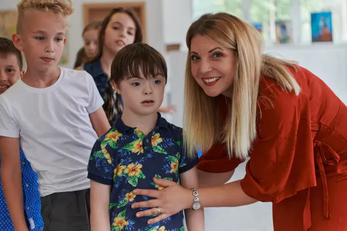 Das Bild zeigt eine Frau in rotem Kleid, die lächelnd neben einem Jungen mit Down-Syndrom steht und ihn sanft am Bauch berührt. Um die beiden herum sind weitere Kinder, die neugierig in die Kamera schauen. Die Szene wirkt freundlich und unterstützt, als ob sie in einer Schule oder Betreuungsumgebung aufgenommen wurde.