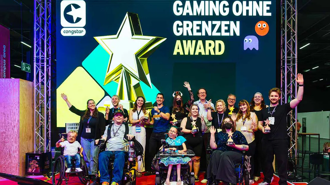 Das Bild zeigt eine große Gruppe von Menschen, darunter mehrere Personen im Rollstuhl, die gemeinsam auf einer Bühne stehen und in die Kamera lächeln. Im Hintergrund leuchtet ein großes Display mit einem goldenen Stern und der Aufschrift „Gaming ohne Grenzen Award“. Viele der Anwesenden halten Trophäen in den Händen und heben jubelnd die Arme.