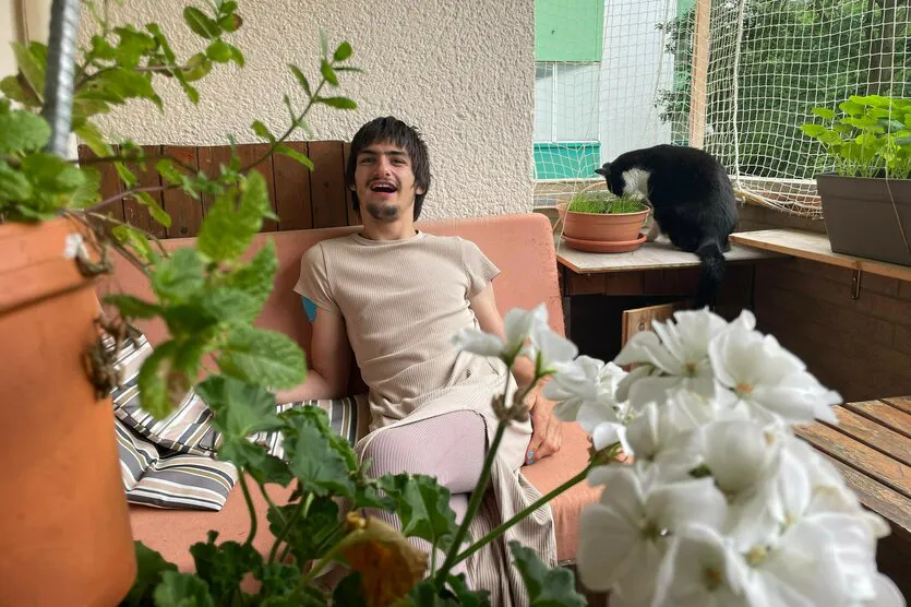 Das Bild zeigt einen jungen Mann, der entspannt auf einem Sofa auf einem Balkon sitzt und fröhlich lacht. Im Vordergrund sind Pflanzen und weiße Blüten zu sehen, während im Hintergrund eine schwarz-weiße Katze aus einem Blumentopf frisst. Der Balkon ist mit weiteren Pflanzkübeln und einem Schutznetz ausgestattet.