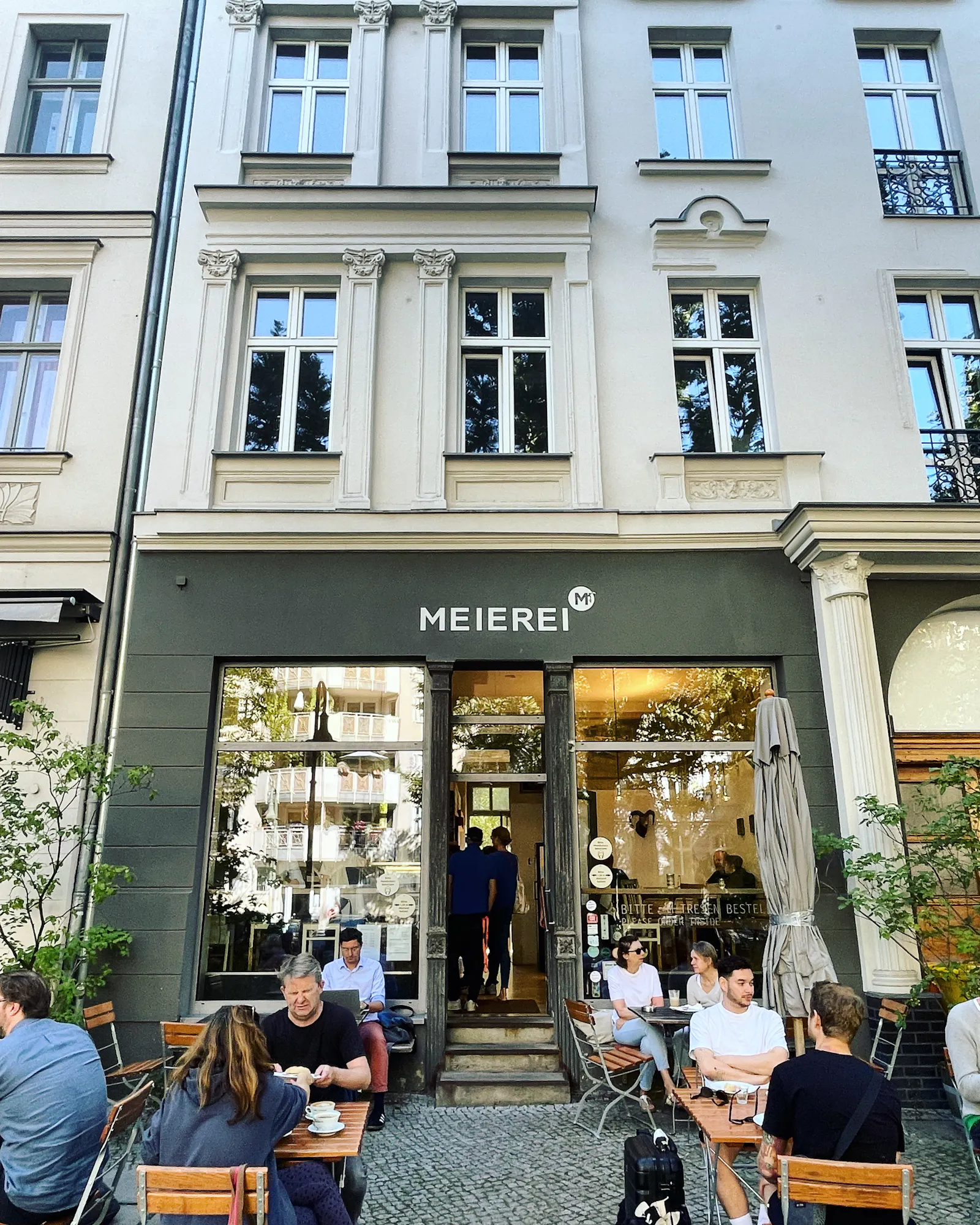 Sommerlich gekleidete Menschen sitzen auf hölzernen Klappstühlen an kleinen Tischchen vor einem Café in einem Gründerzeithaus. Innen sieht man warme Beleuchtung und weitere Menschen.