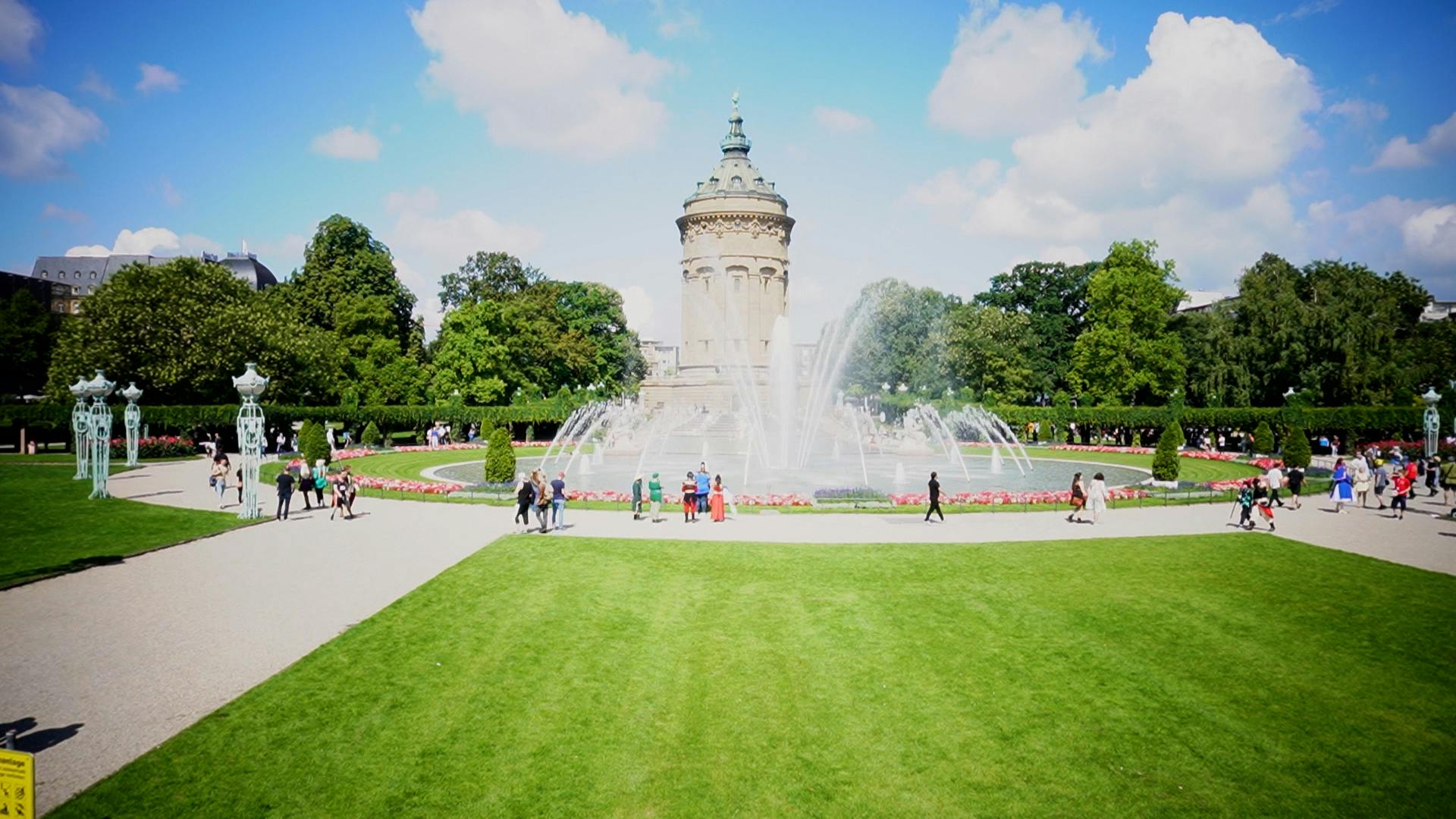 Eine Sommeransicht des Mannheimer Wasserturms im Rosengarten: Der historische, säulenverzierte Turm mit kupfergrüner Kuppel steht im Hintergrund vor blauem Himmel mit weißen Wolken, umgeben von einem großen Springbrunnen. Davor erstreckt sich eine gepflegte Grünanlage mit Wegen, Blumenbeeten und mehreren spazierenden Besuchern, manche davon offenbar in Cosplay-Kostümen.