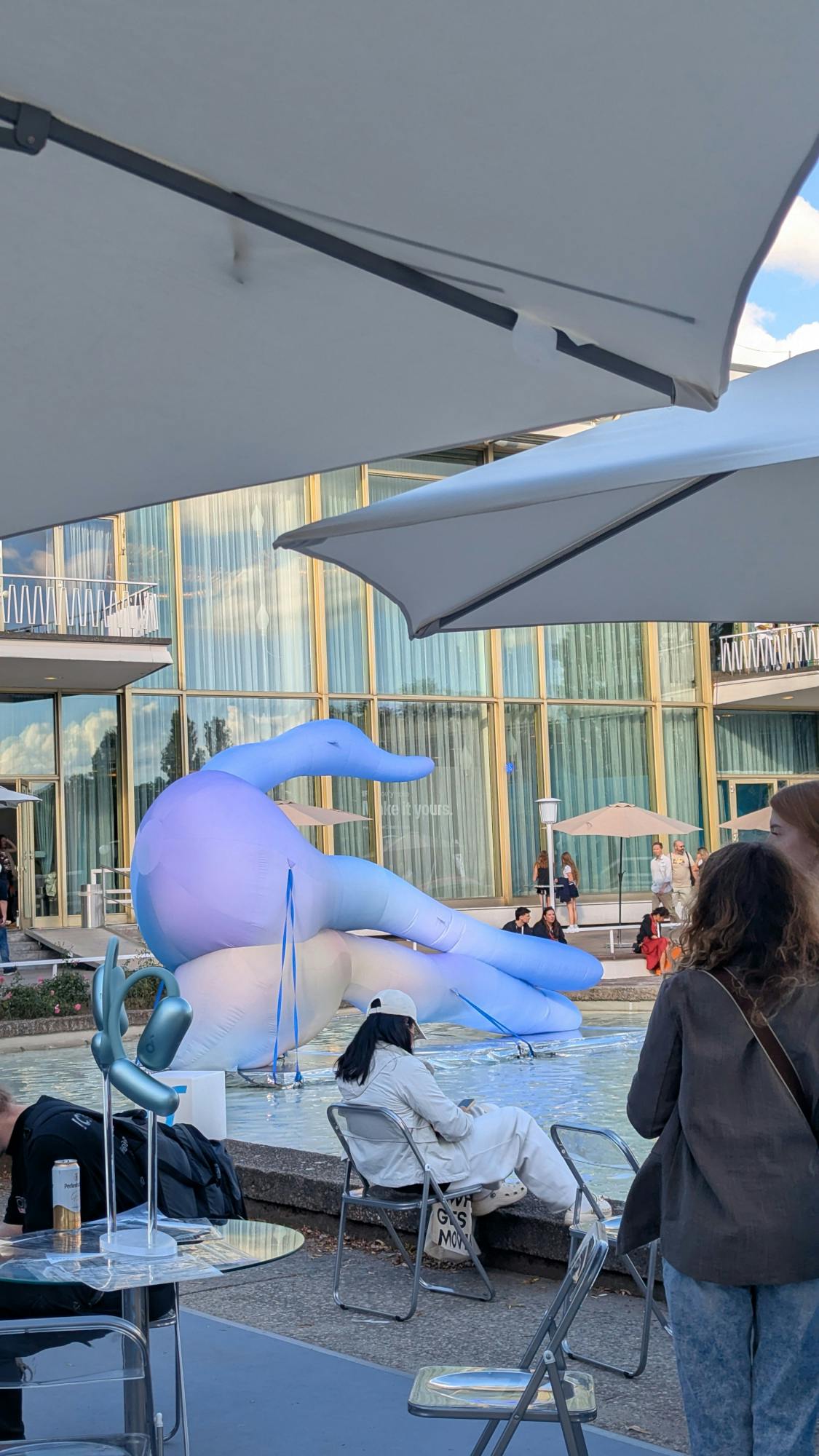 Ein aufgeblasenes, blau-violett-gelb changierendes Objekt auf einem silbrigen Podest in einem Teich, hinter den Sonnenschirmen eines Cafés, vor einer hohen Glasfassade, die nach Fünfzigerjahren aussieht.