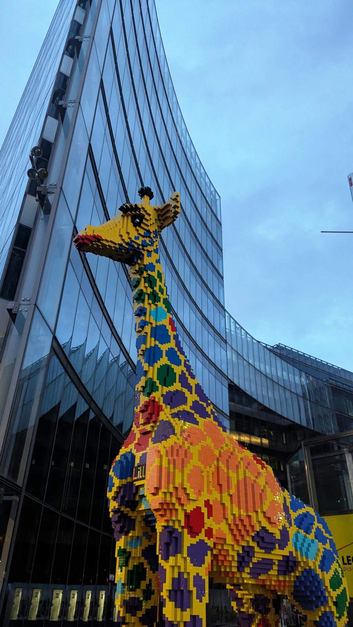 Eine riesige Legogiraffe vor einer geschwungenen, spiegelverglasten Hochhausfassade vor bewölktem Abendhimmel.