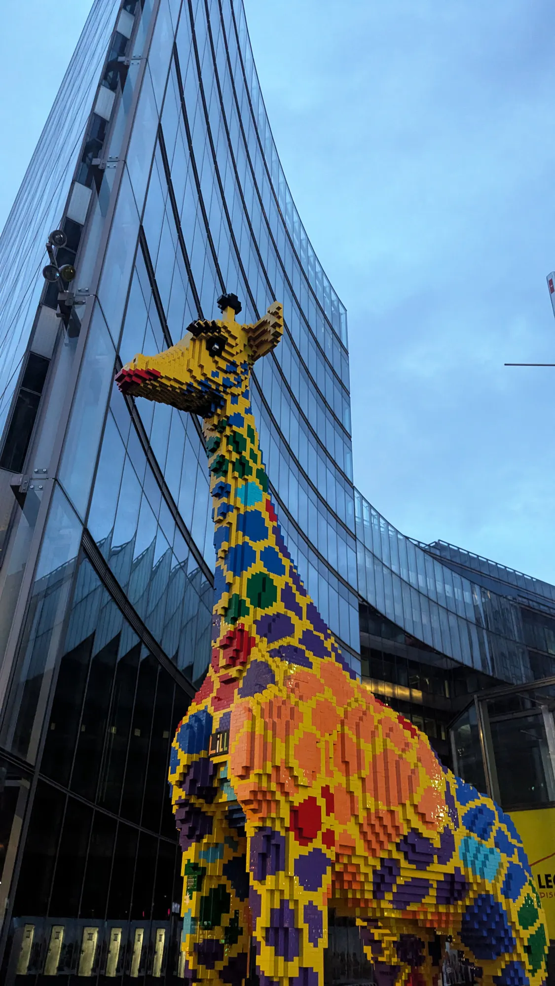 Eine riesige Legogiraffe vor einer geschwungenen, spiegelverglasten Hochhausfassade vor bewölktem Abendhimmel.
