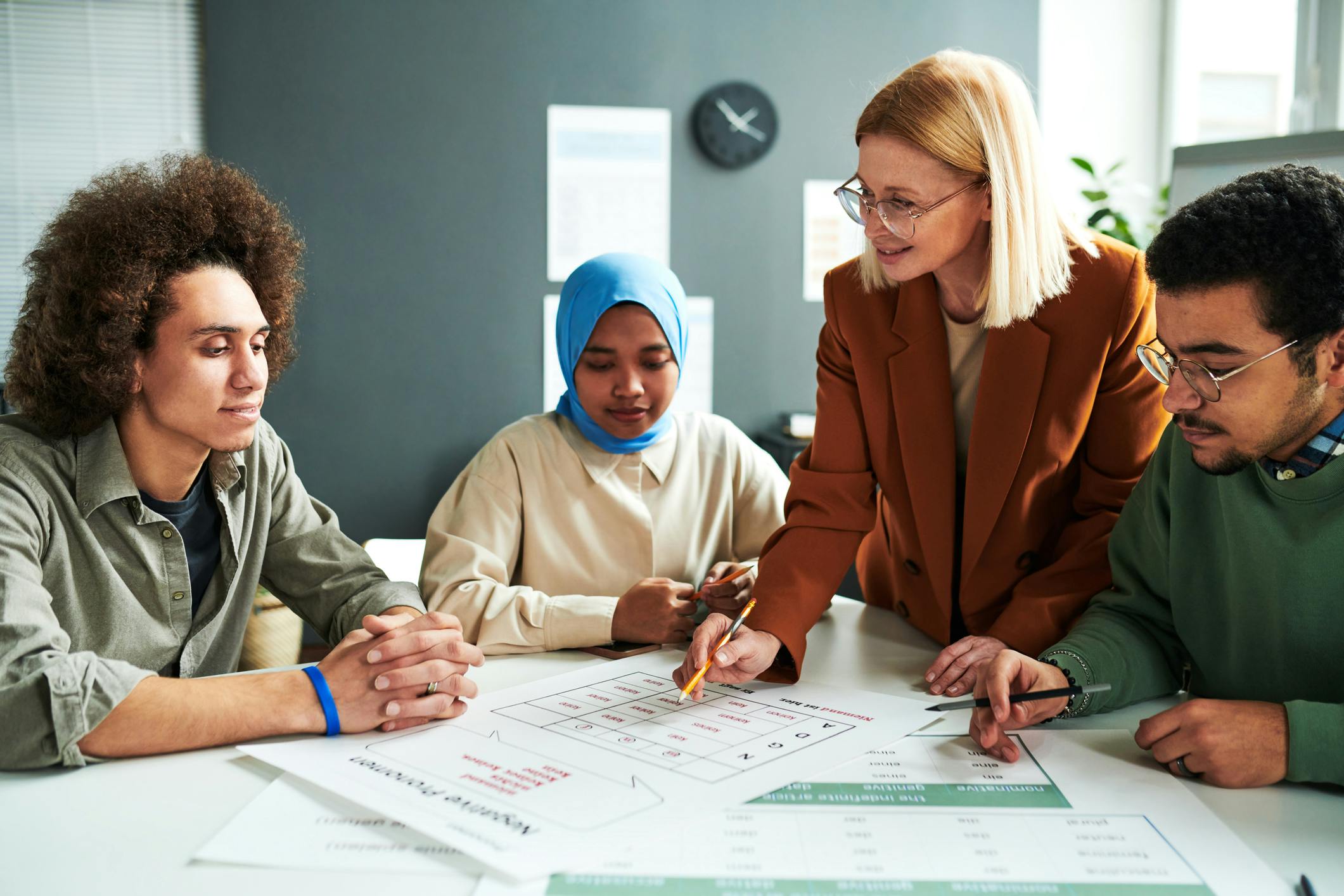 Dieses Foto zeigt eine Gruppe erwachsener Kursteilnehmer:innen und eine Lehrerin, die gemeinsam an einem Tisch sitzen und über Arbeitsblättern einen Sprachkurs-Inhalt besprechen.