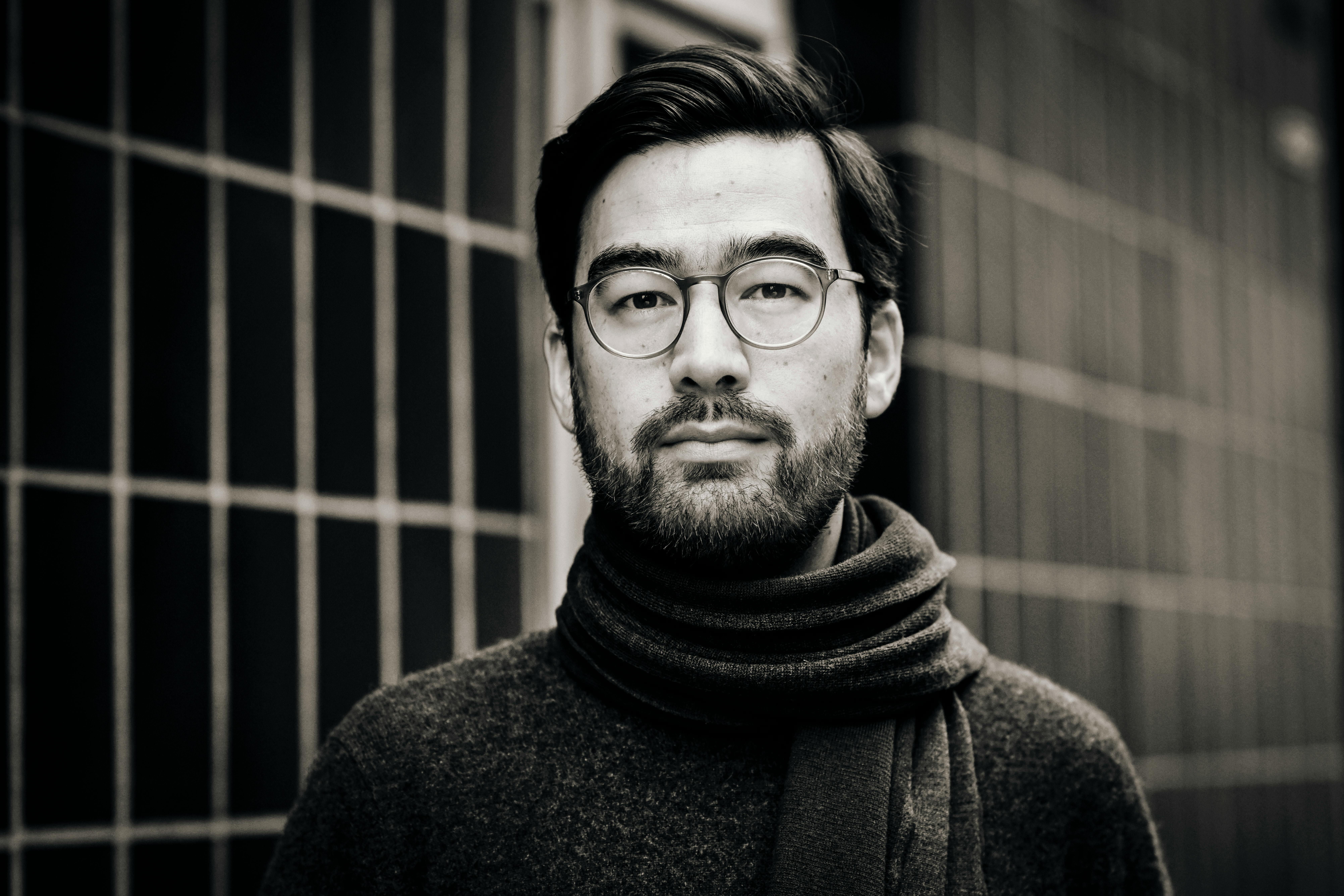 Yannic Han Biao Federer  trägt einen dunklen Wollpullover und einen Schal. Er schaut ernst in die Kamera, hinter ihm ist ein Gitter zu sehen. Das Bild ist in Schwarz-Weiß.