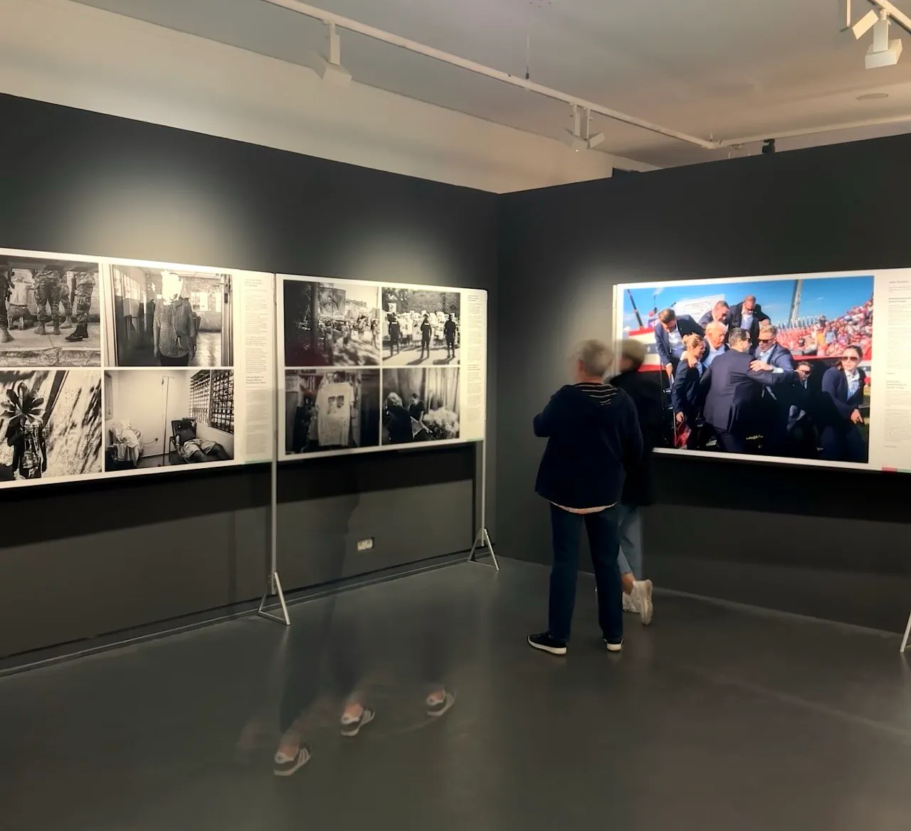 Foto aus der Ausstellung: Zwei Stellwände voller Fotos, davor verschwommen Personen.