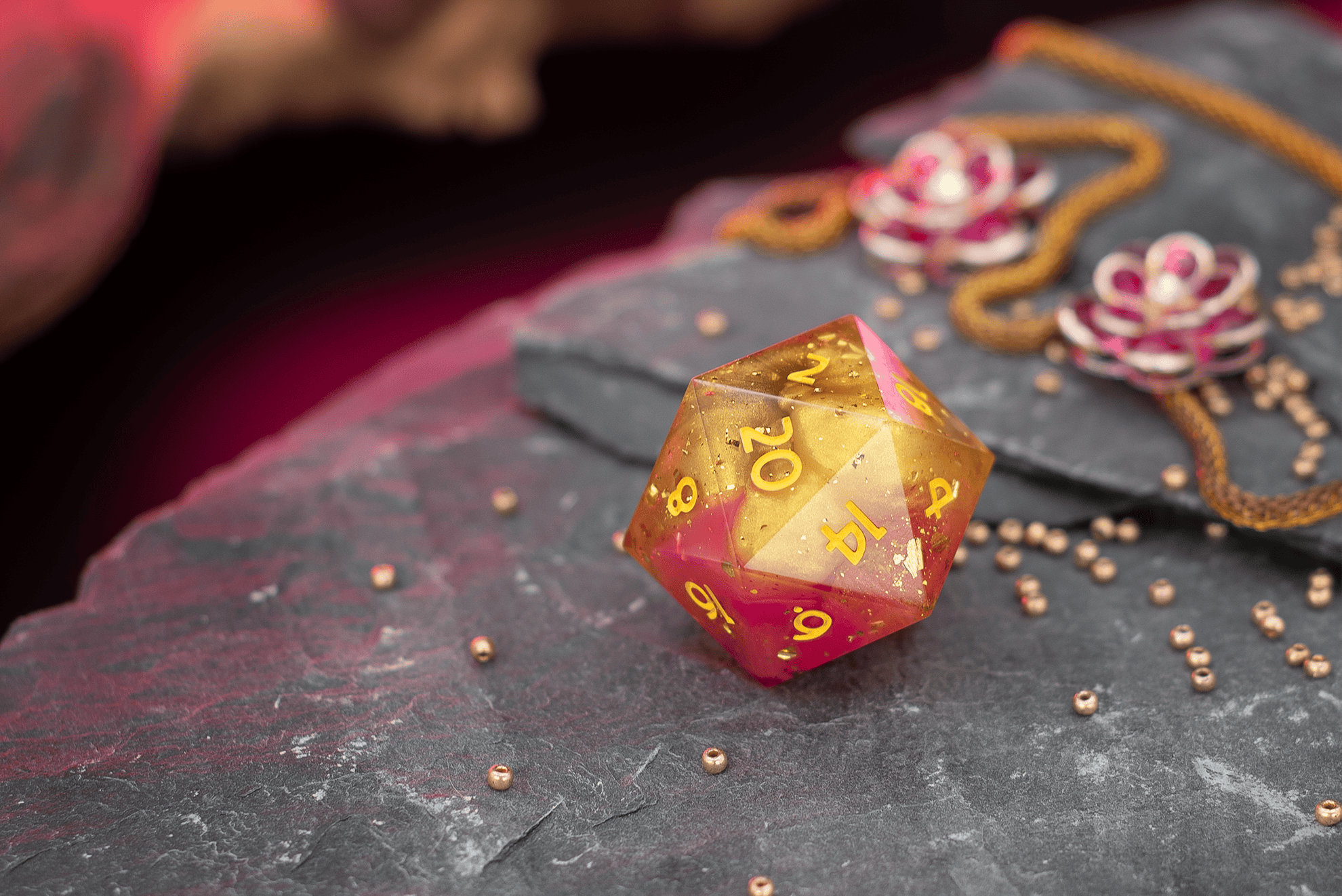 Rot-goldener, glitzernder W20-Würfel mit goldenen Zahlen liegt auf dunklem Schieferstein, umgeben von kleinen goldenen Perlen; im Hintergrund unscharf Schmuckelemente und warme pink-goldene Beleuchtung.