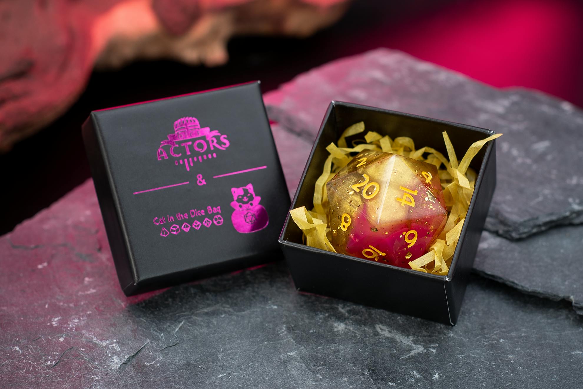 Schwarze Geschenkbox mit pinkfarbenem „Dice Actors & Cat in the Dice Bag“-Logo, daneben eine geöffnete Box mit einem großen, rot-goldenen, glitzernden W20-Würfel auf gelbem Papierfüllmaterial, fotografiert auf dunklem Steinuntergrund mit pinkfarbener Beleuchtung.