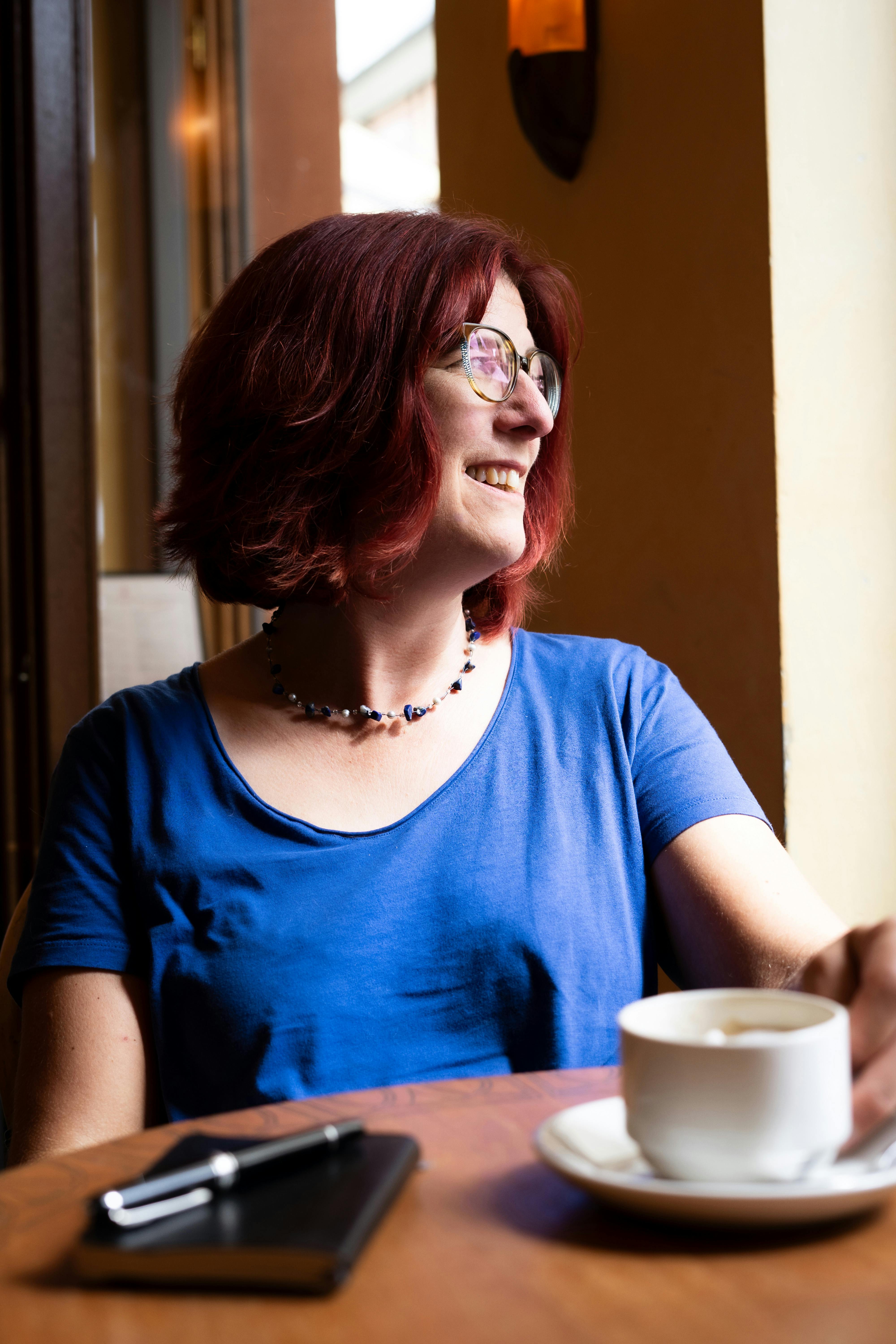 Diana Menschig, weiß, rot gefärbte Haare, im Halbprofil nach rechts schauend, gekleidet mit einem blauen T-Shirt. Vor ihr auf dem Tisch eine weiße Tasse mit Untertasse, ein schwarzes Notizbuch und ein schwarzer Füller