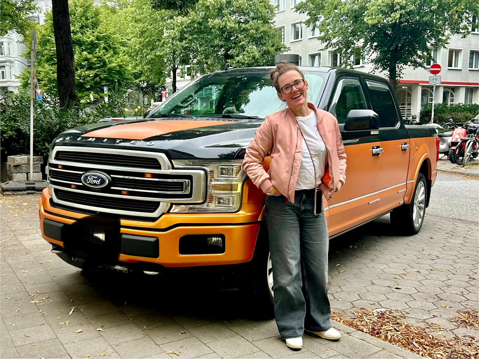Eine Frau steht lächelnd und mit den Händen in den Taschen vor einem riesigen, zweifarbig lackierten Ford-Pick-up, der orange und schwarz gestaltet ist. Er parkt auf dem Gehweg.
Die Szene spielt auf einem baumbestandenen Gehweg in einer Wohngegend, im Hintergrund sind mehrstöckige Häuser und geparkte Fahrzeuge zu sehen.