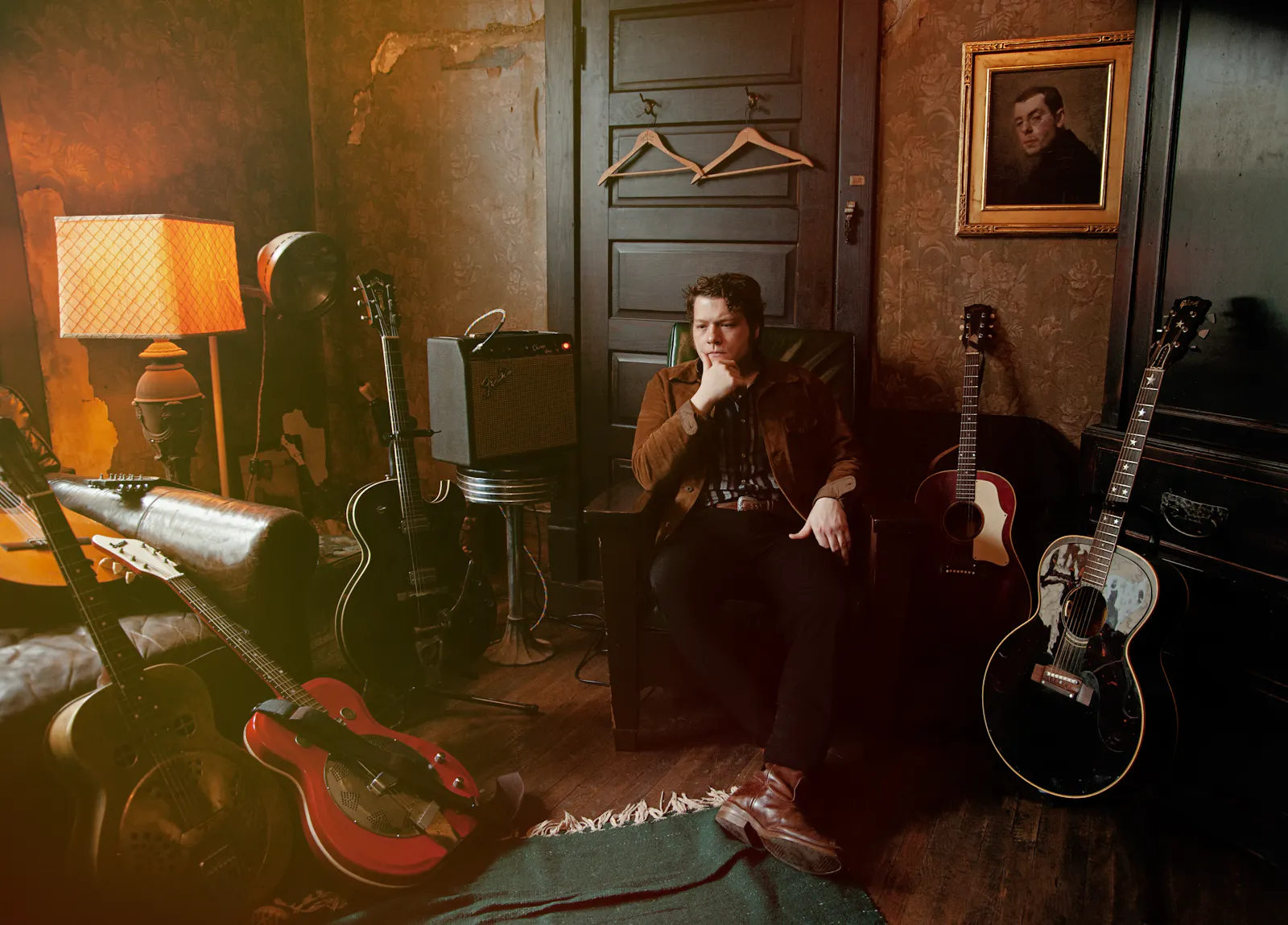 Foto eines jungen Mannes, der umgeben von mehreren Gitarren nachdenklich in altmodischer Kleidung in einer altmodischen Kulisse sitzt