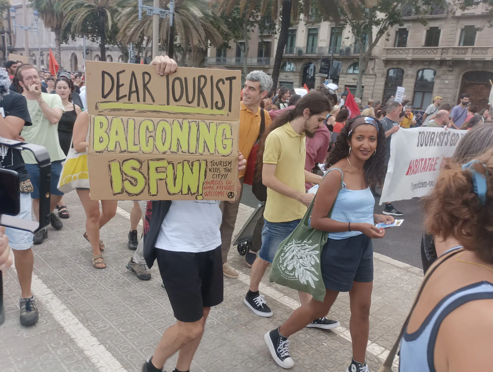 Auf einer Demonstration gegen Overtourism in Barcelona im Juli 2024 hält ein Mann ein selbstgemaltes Plakat mit der Aufschrift "Dear tourist, balconing is fun" in die Kamera.