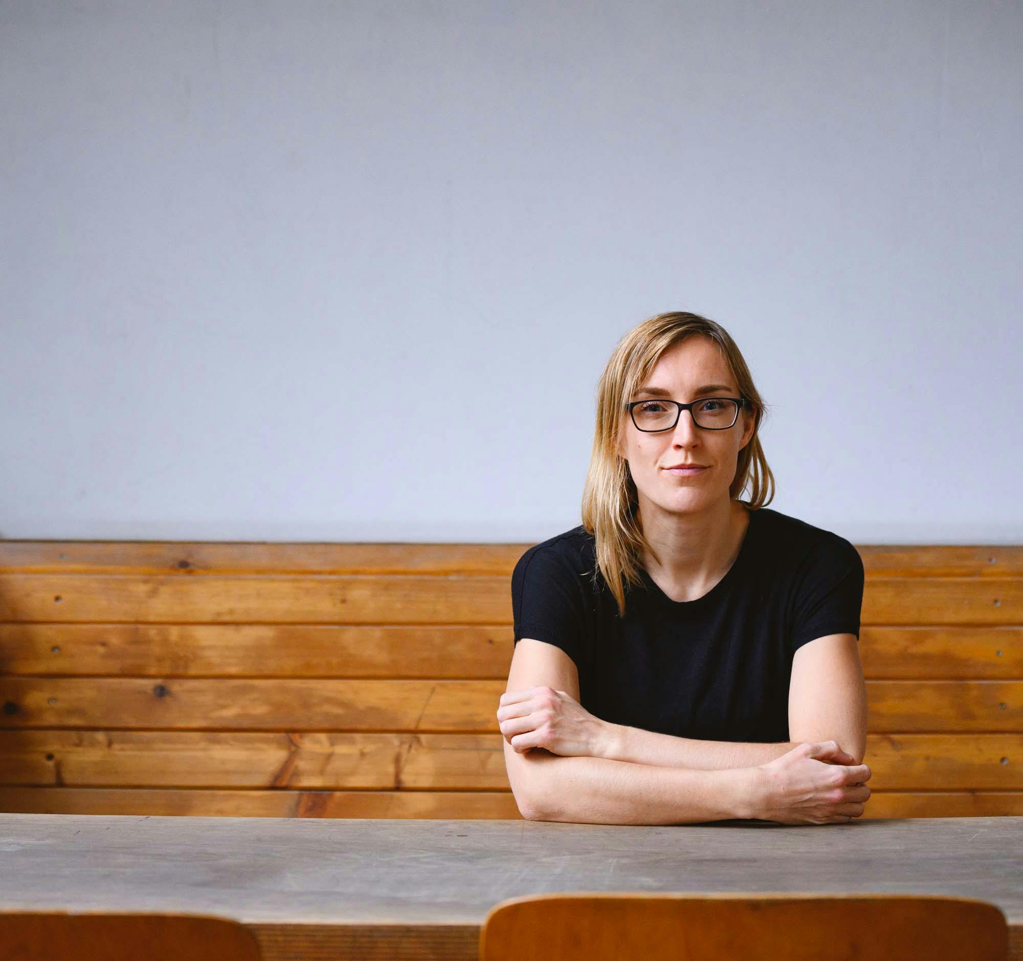 Juliane Fritz, eine junge Frau im schwarzen Shirt mit langen blonden Haaren und einer schwarzen Brille sitzt an einem Tisch mit verschränkten Armen
