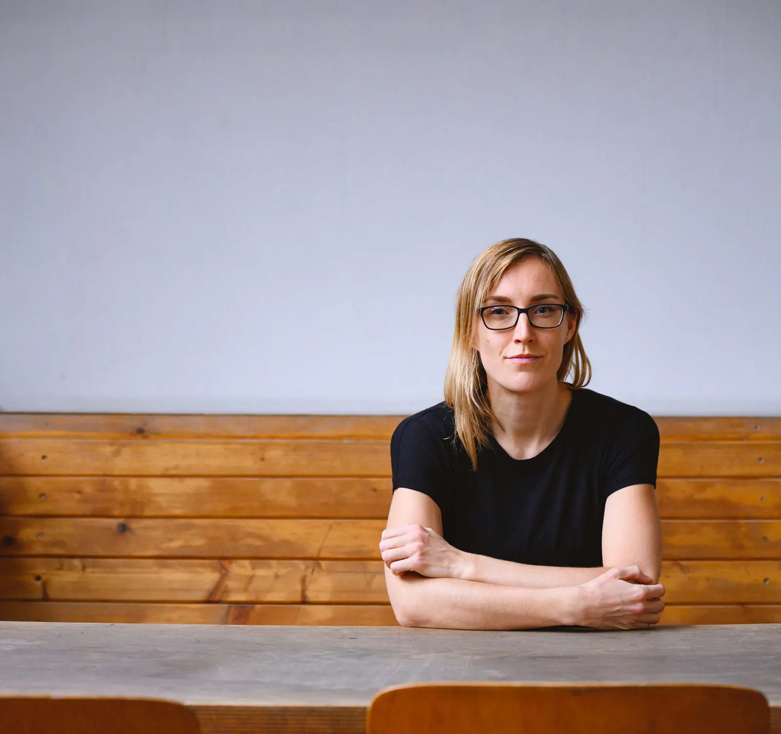 Juliane Fritz, eine junge Frau im schwarzen Shirt mit langen blonden Haaren und einer schwarzen Brille sitzt an einem Tisch mit verschränkten Armen