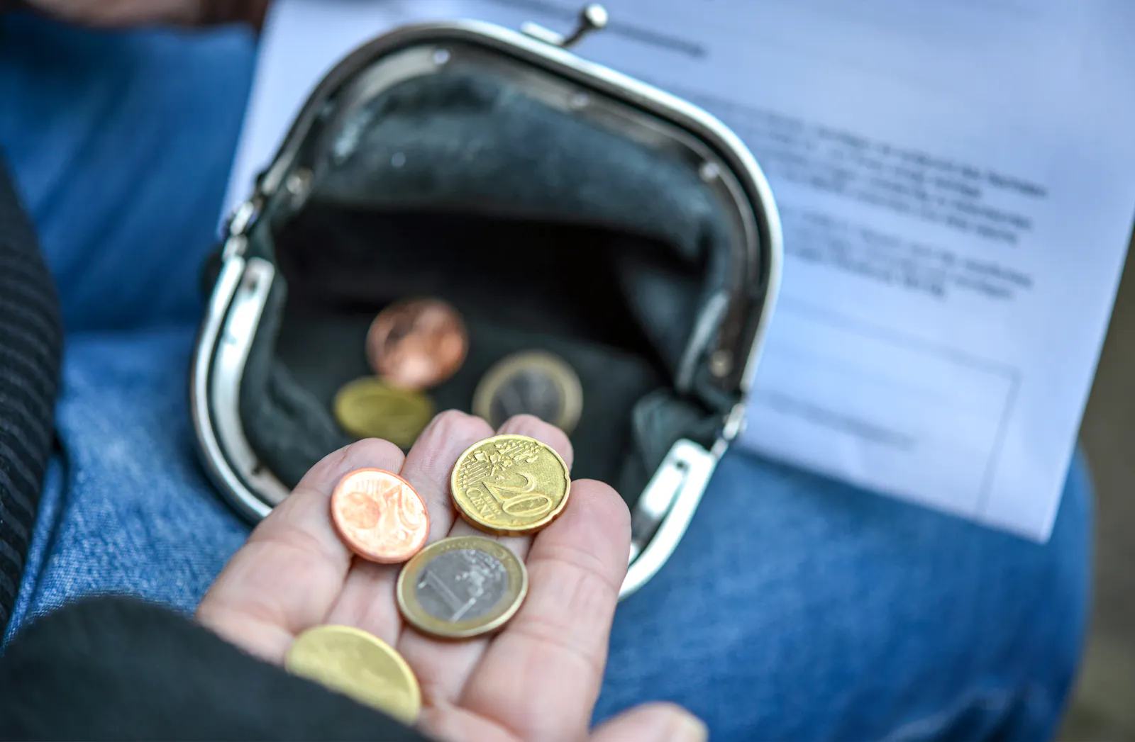 Das Bild zeigt eine Hand mit ein paar Euro-Münzen vor einer leeren Geldbörse. Im Hintergrund ist ein unscharfes Dokument.