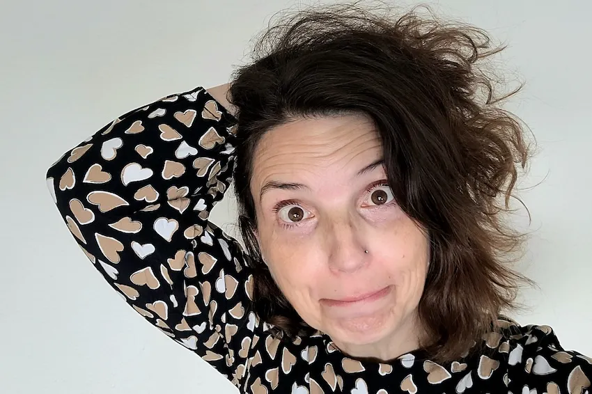 Christine Piontek im schwarzen Oberteil mit weißen und beigen Herzen schaut erstaunt und fährt sich durch die Haare.
