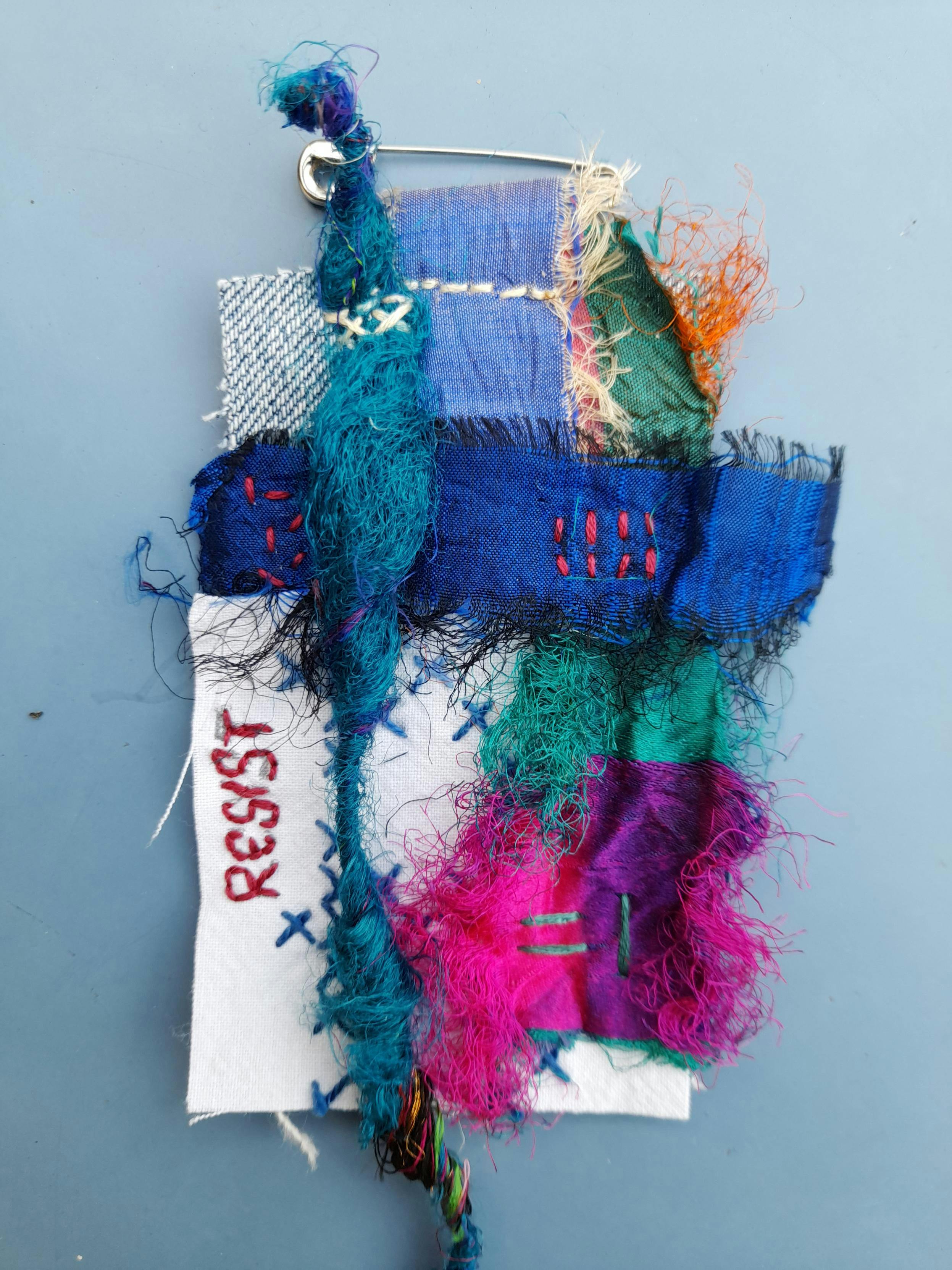 A multicoloured collage with textile scraps, wild intuitive stitchingand the embroidered word RESIST.