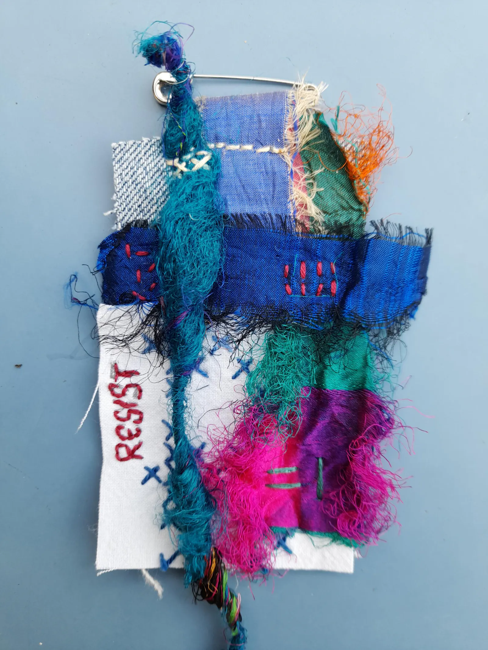 A multicoloured collage with textile scraps, wild intuitive stitchingand the embroidered word RESIST.