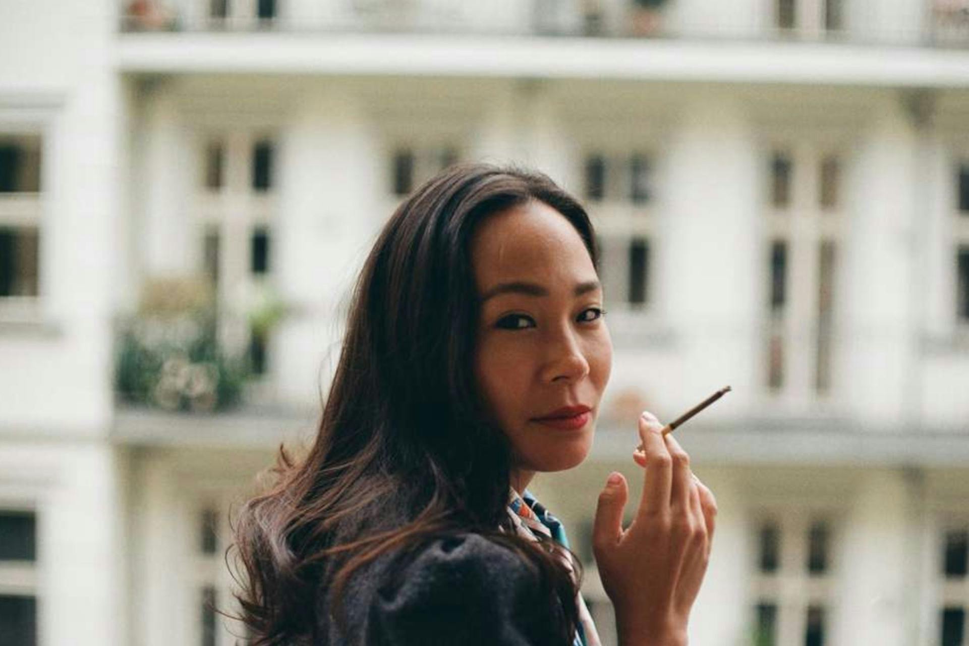 Ta-Som Helena Yun steht auf einem Balkon, hinter ihr ist eine Häuserfassade zu sehen. Sie trägt ein dunkles Oberteil und eine dünne Zigarette in der Hand.