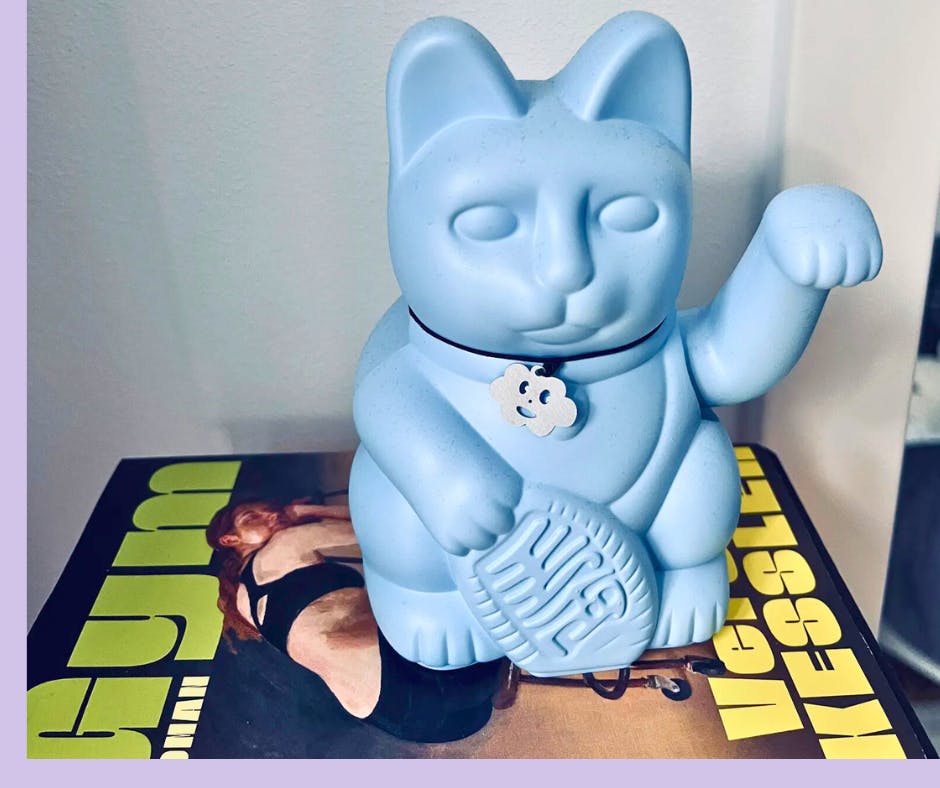 Eine hellblaue Winkekatze, die mit dem linken Arm winkt, steht auf einem Stapel Bücher