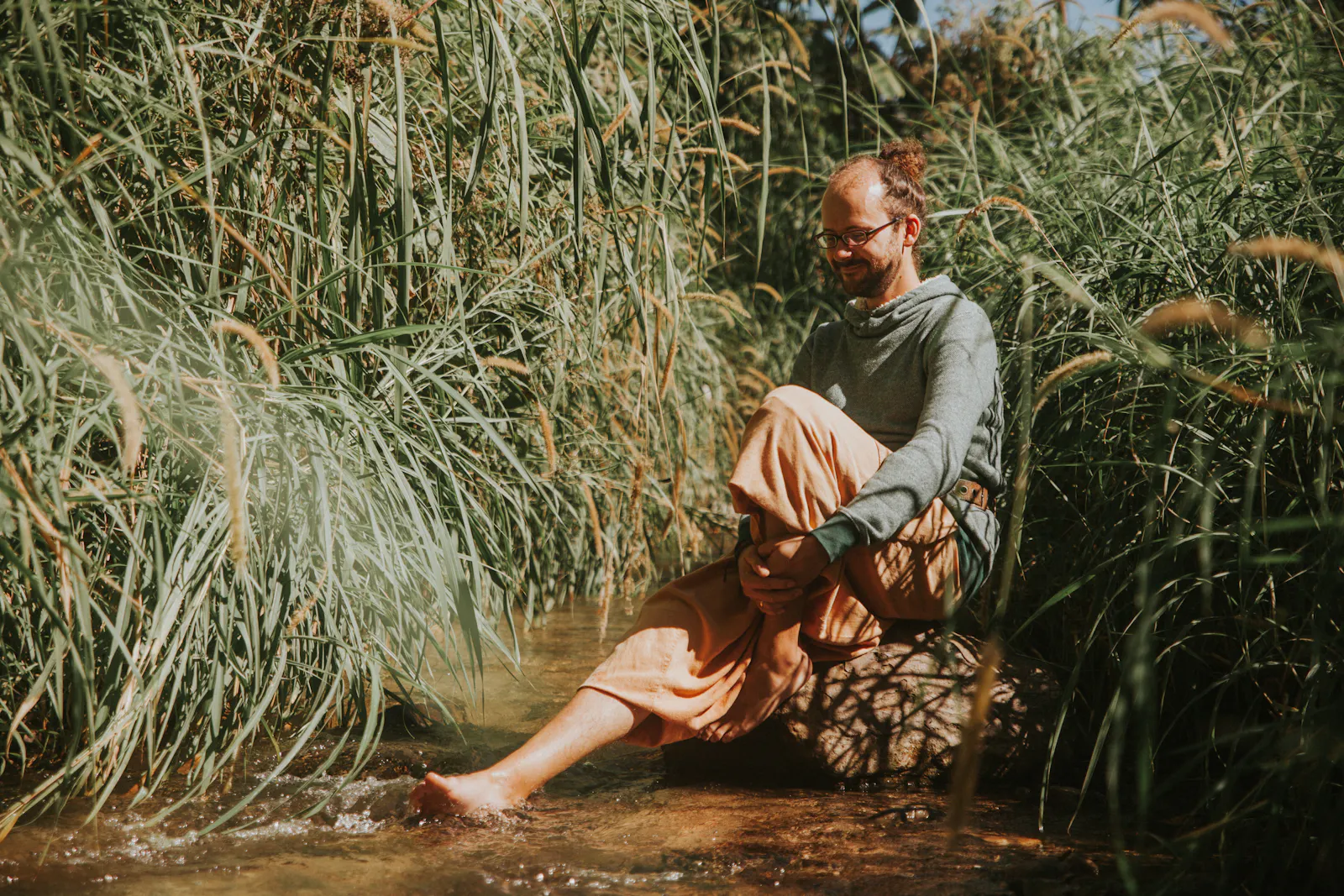 Olaf sitzt mit nackten Füßen auf einem Stein im Flussbett und hält die Füße ins Wasser. Um ihn herum grüner Farn.