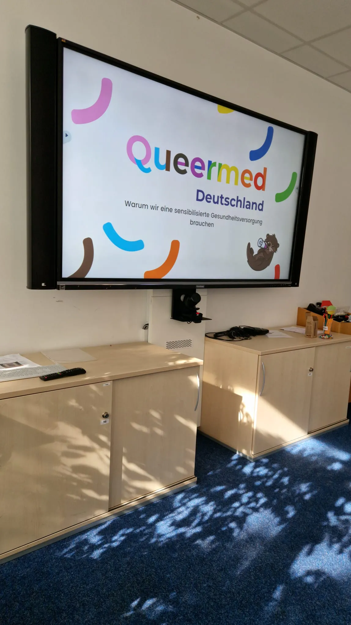 An einem großen Bildschirm in einem Raum, der an einer Wand befestigt ist, befindet sich ein Slide mit dem Logo von Queermed Deutschland.
Unter dem Bildschirm an der Wand befinden sich mehrere kleine Schränke.