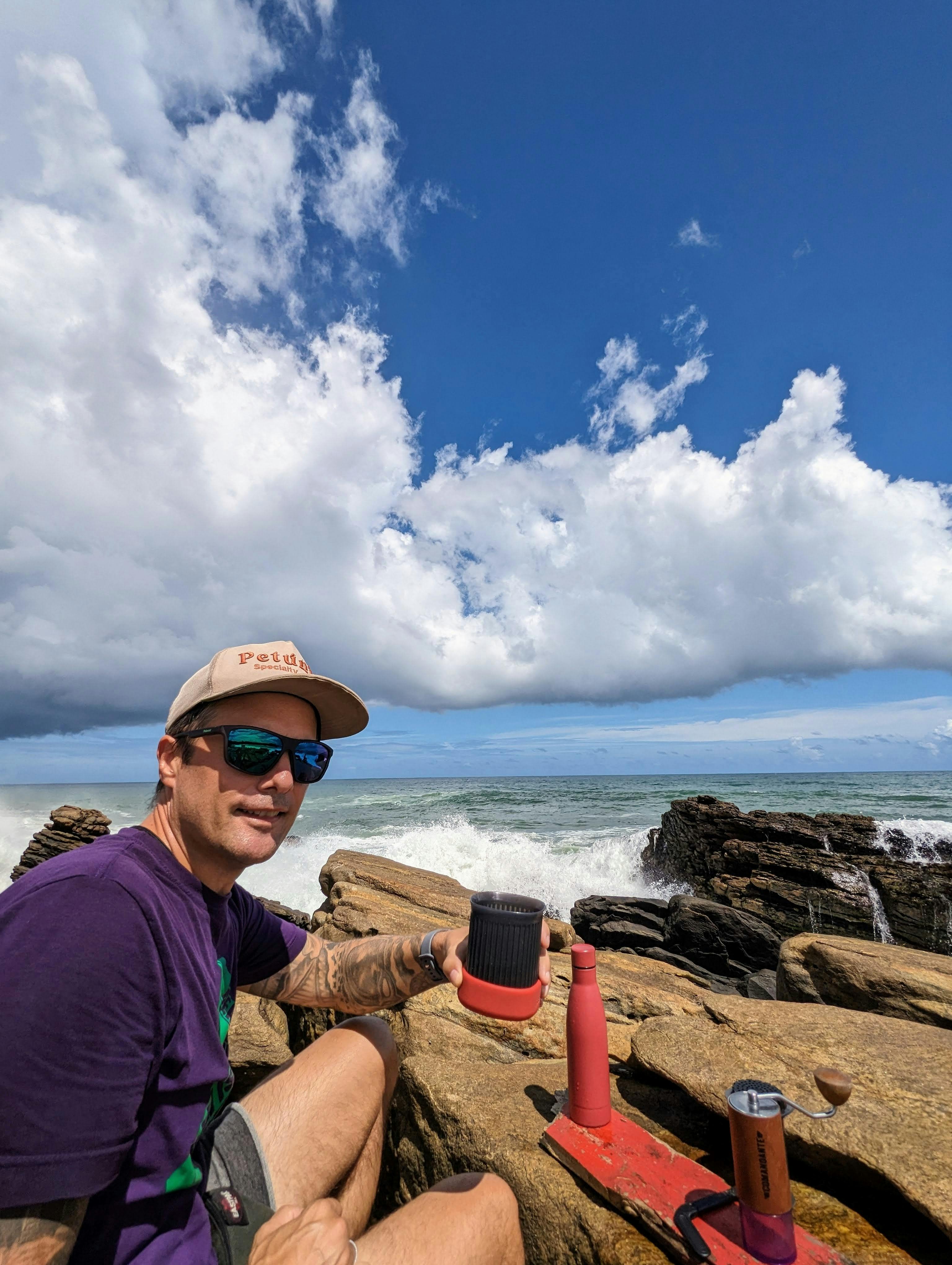 Daniel von Coffeeteller sitzt im lilafarbenen T-Shirt, mit Sonnenbrille und Cap auf der dunklen Felsküste von Sri Lanka. Direkt hinter ihm bricht sich die weiße Gischt des Indischen Ozeans an den Steinen. Vor ihm auf den Felsen steht sein kompaktes Reise-Equipment: eine Handmühle und ein Becher. Er blickt lächelnd in die Kamera und hält seine Kaffeetasse hoch. Über ihm erstreckt sich ein tiefblauer Himmel mit markanten, weißen Schönwetterwolken.