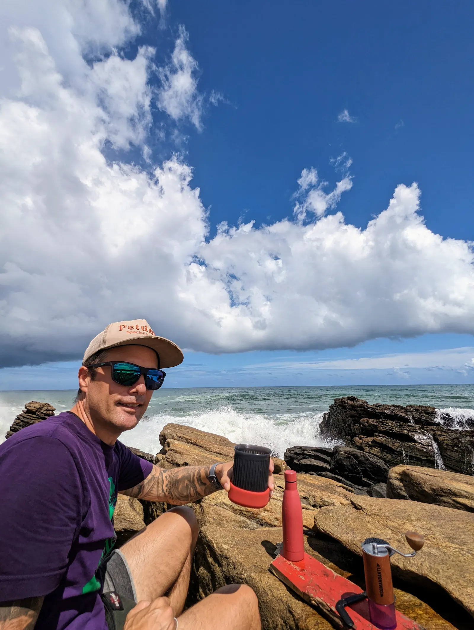 Daniel von Coffeeteller sitzt im lilafarbenen T-Shirt, mit Sonnenbrille und Cap auf der dunklen Felsküste von Sri Lanka. Direkt hinter ihm bricht sich die weiße Gischt des Indischen Ozeans an den Steinen. Vor ihm auf den Felsen steht sein kompaktes Reise-Equipment: eine Handmühle und ein Becher. Er blickt lächelnd in die Kamera und hält seine Kaffeetasse hoch. Über ihm erstreckt sich ein tiefblauer Himmel mit markanten, weißen Schönwetterwolken.
