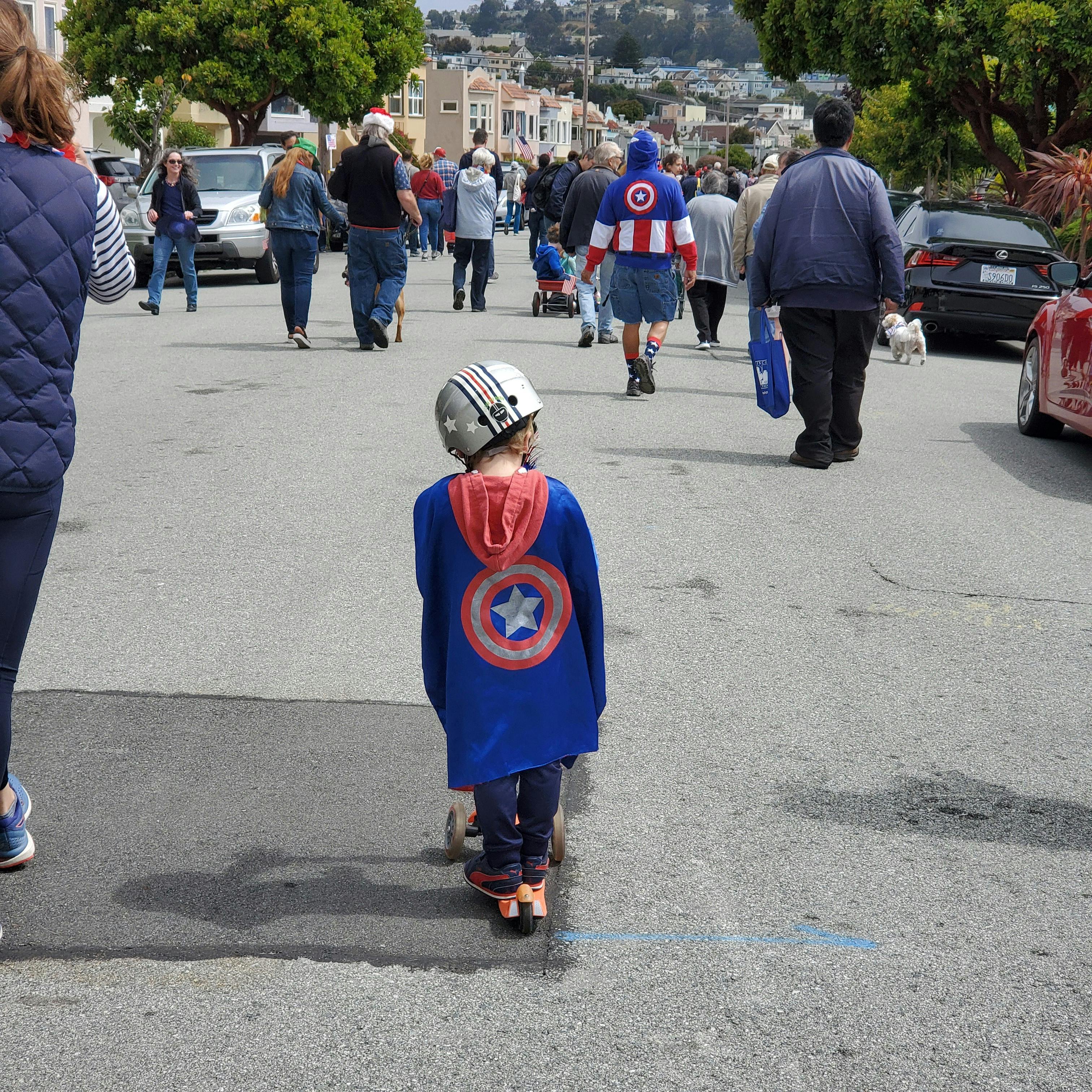 Foto einer Gruppe von Menschen, die in losem Zusammenhang eine Straße in San Francisco entlanggeht. Alle sind von hinten zu sehen im Mittelpunkt ist der Rücken eines Jungen auf einem Roller zu sehen, der ein Captain America Cape und Helm trägt. Beides hängt etwas schief an ihm und er scheint zu Boden zu blicken