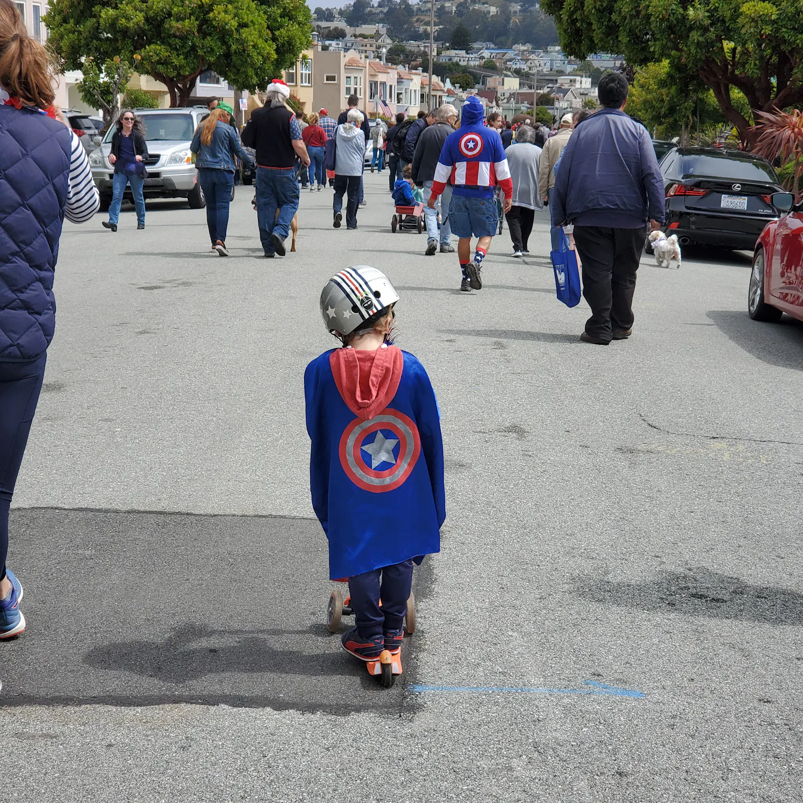 Foto einer Gruppe von Menschen, die in losem Zusammenhang eine Straße in San Francisco entlanggeht. Alle sind von hinten zu sehen im Mittelpunkt ist der Rücken eines Jungen auf einem Roller zu sehen, der ein Captain America Cape und Helm trägt. Beides hängt etwas schief an ihm und er scheint zu Boden zu blicken