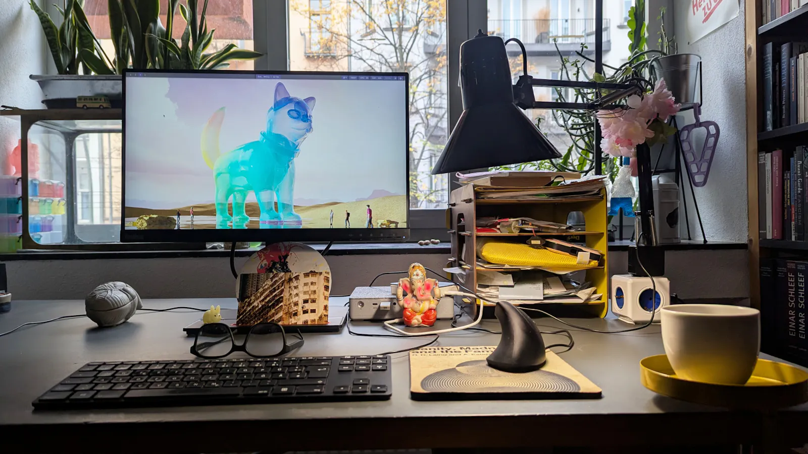 Conputerbildschirm mit einer KI-generierten Katzen-Sphinx aus Buntglas als Bildschirmhintergrund, Schreibtischlampe, Laffeetasse, ein kleiner Ganesha, hinten auf der Fensterbank zwei Erdnüsse.