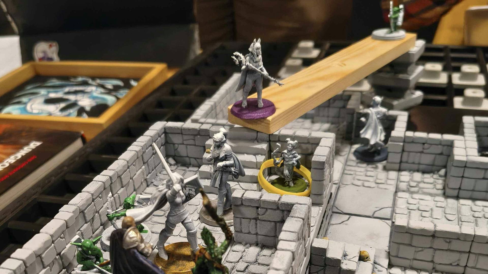 Eine Kampfsituation auf einer Battlemap mit zum Teil unbemalten Miniaturen von Helden und Goblins. Die Karte scheint ein steinerner Dungeon zu sein mit Treppen, verschieden hohen Ebenen und einer Behelfsbrücke aus Holz.