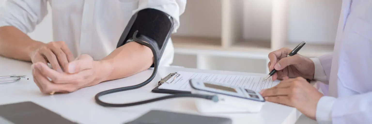 Blutdruckmessung beim Arzt: Mann in weißem Shirt sitzt am Tisch, hat eine Blutdruckmanschette am Arm. Daneben ein Arzt, der mit einem Stift die Werte auf eine Kladde schreibt.