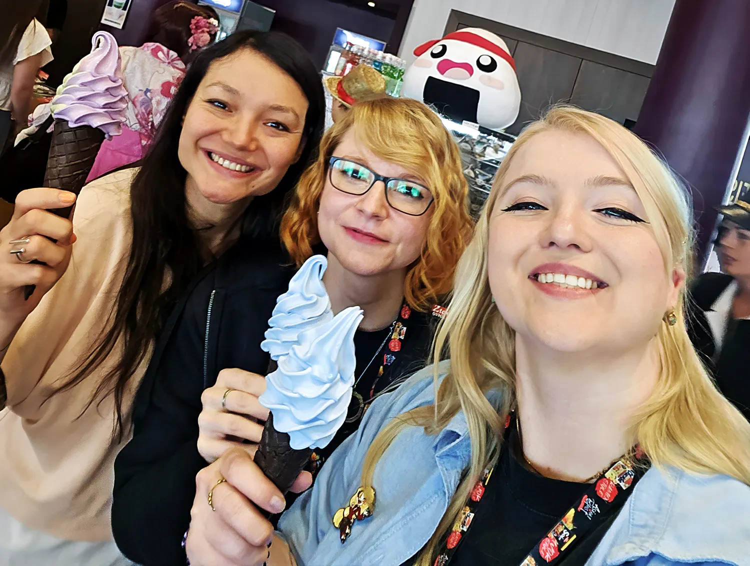 Rubi, Stephie und Rieke lächeln in die Kamera und halten Eiswaffeln mit lilafarbenem und hellblauem Softeis in der Hand.