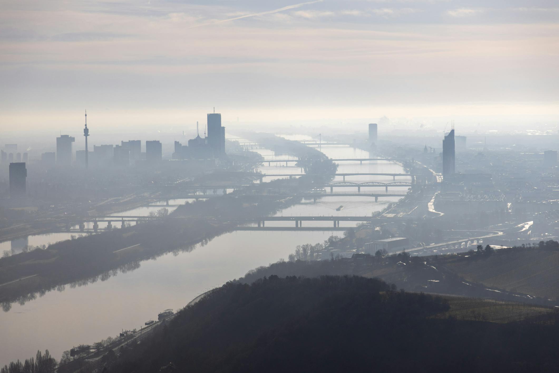 Das Foto zeigt eine Luftaufnahme von Wien an einem nebligen Tag. Die Donau fließt durch die Bildmitte, mehrere Brücken verbinden die beiden Ufer. Links im Bild ist der Donauturm zu sehen. Hochhäuser und Gebäude sind im dichten Nebel nur schemenhaft zu erkennen. Der Himmel ist grau und trüb.