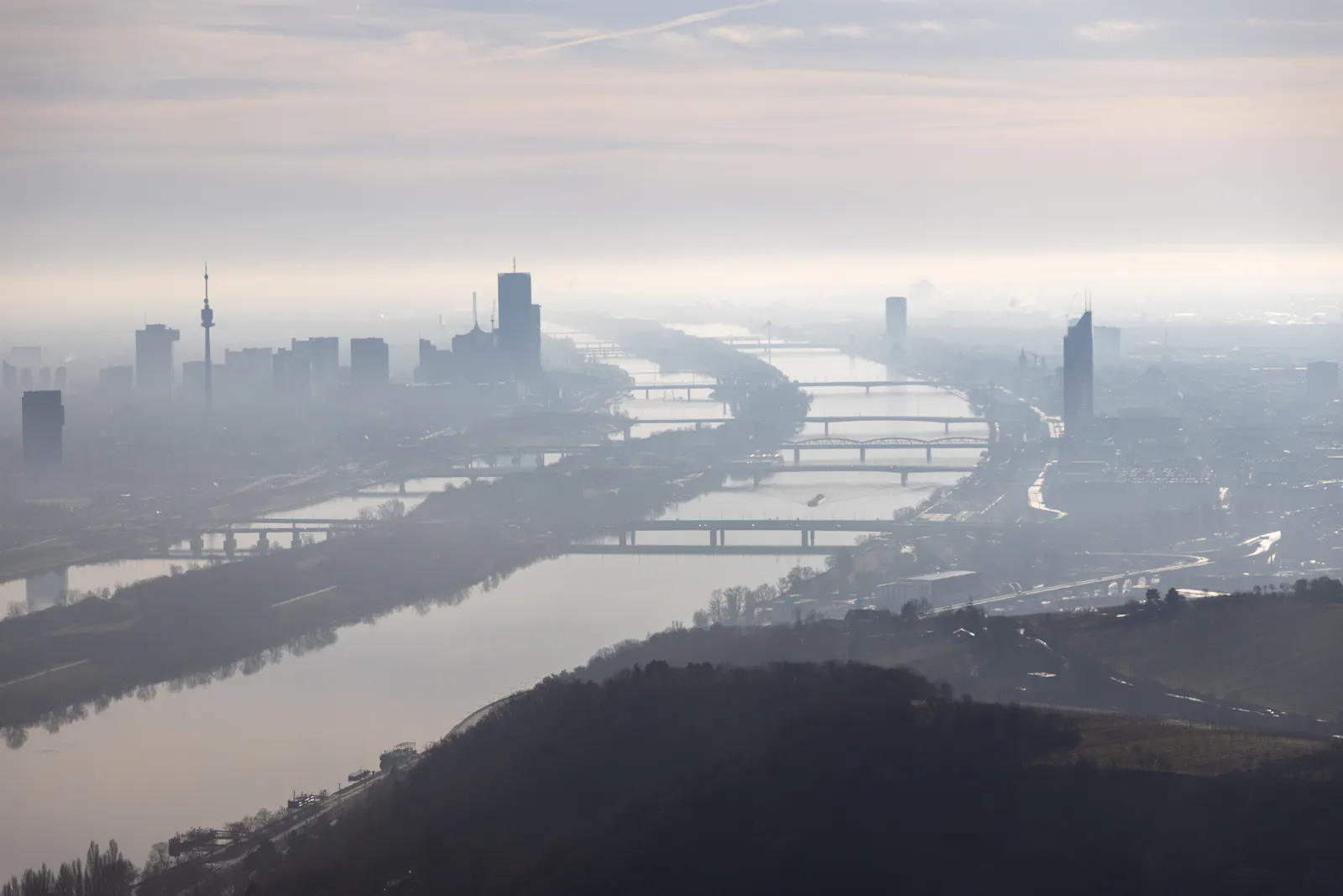 Das Foto zeigt eine Luftaufnahme von Wien an einem nebligen Tag. Die Donau fließt durch die Bildmitte, mehrere Brücken verbinden die beiden Ufer. Links im Bild ist der Donauturm zu sehen. Hochhäuser und Gebäude sind im dichten Nebel nur schemenhaft zu erkennen. Der Himmel ist grau und trüb.