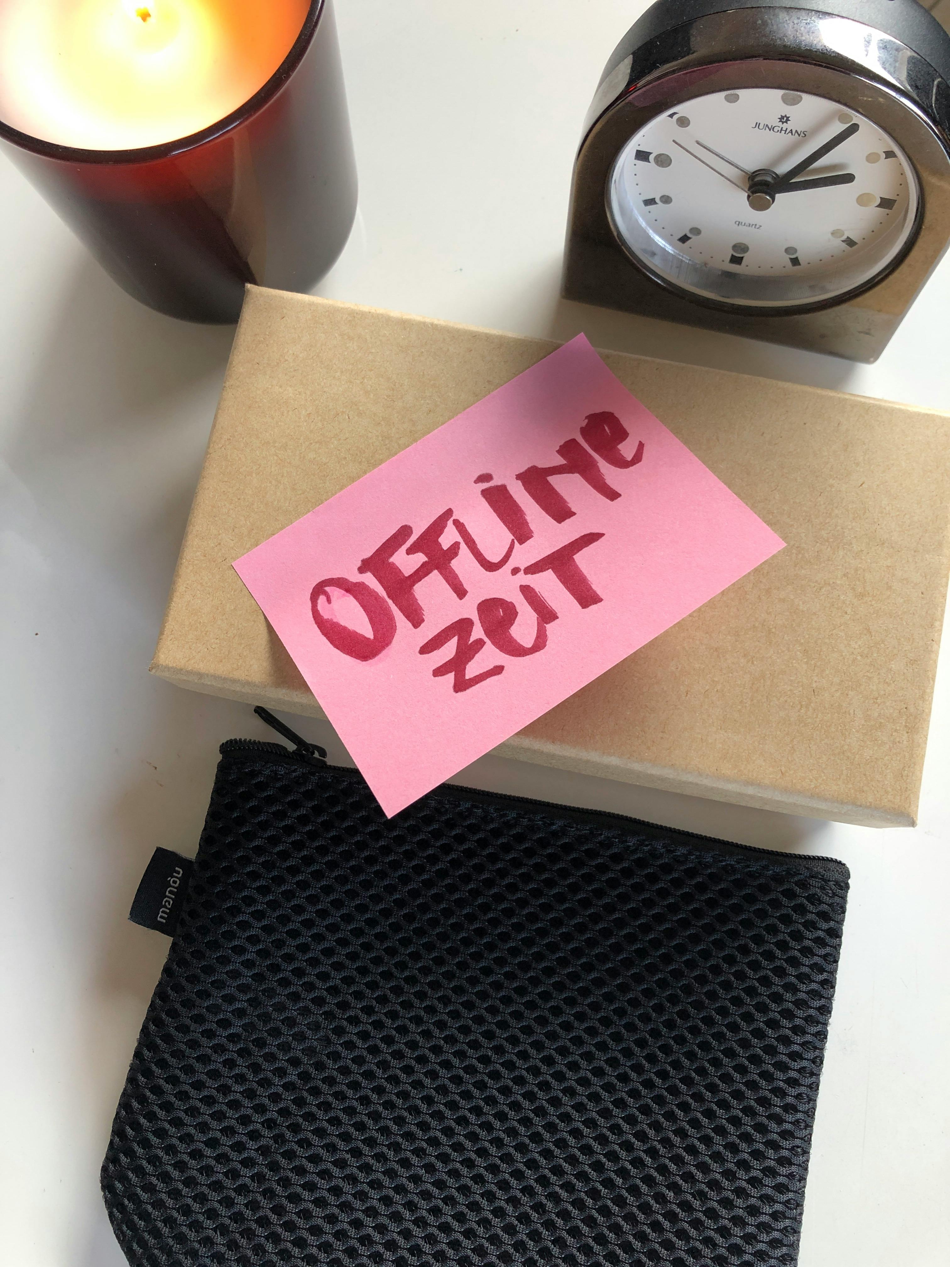 Ein Windlicht, ein analoger Wecker, eine beige Schachtel auf der ein rosa Post-it klebt, auf dem "Offline-Zeit" steht. Darunter ein schwarzes Etui